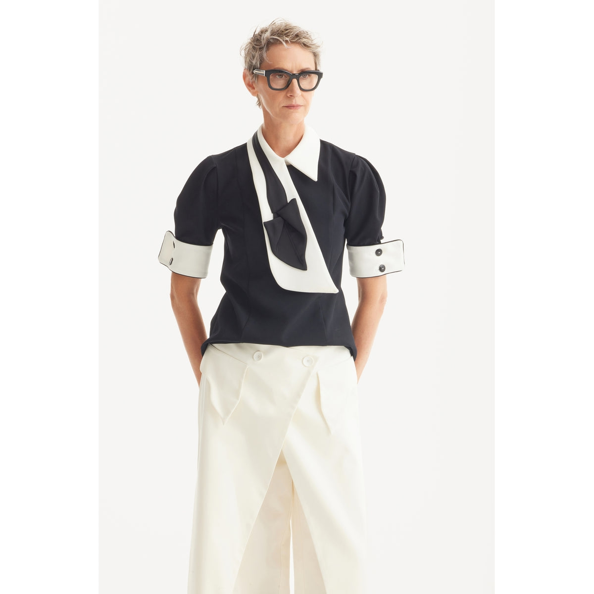 A person with short gray hair and black glasses wears the JULIA ALLERT Statement Blouse With White Collar And Cuffs - Black, paired with wide white high-waisted pants. They stand against a plain white background, hands in pockets.