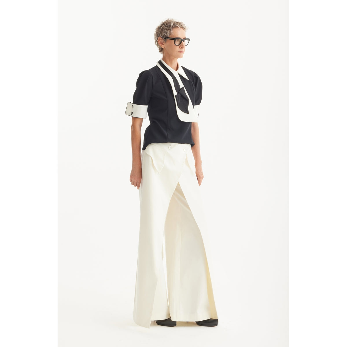 A person with short, curly hair and glasses wears the JULIA ALLERT Statement Blouse With White Collar And Cuffs - Black, paired with wide-legged, high-waisted white pants, standing against a plain white background.