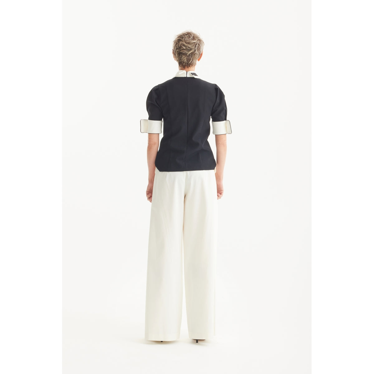 A person with short hair, seen from behind, wears the JULIA ALLERT Statement Blouse With White Collar And Cuffs in black and wide-legged white trousers against a plain white background.