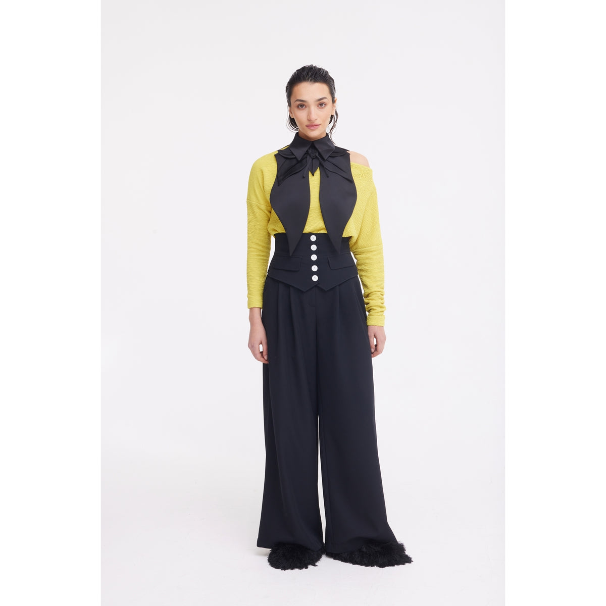 Against a white backdrop, a person wears a yellow long-sleeve top, black bow-tie vest, JULIA ALLERT's Buttoned Corset-Waist Black Tailored Trousers, and black shoes with furry details.