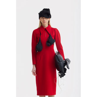A woman in a JULIA ALLERT Solid Formal Midi Dress in red stands against a white background, accessorized with a black sculptural hat, abstract necklace, and holding a large black bag featuring loose threads and attached shapes.