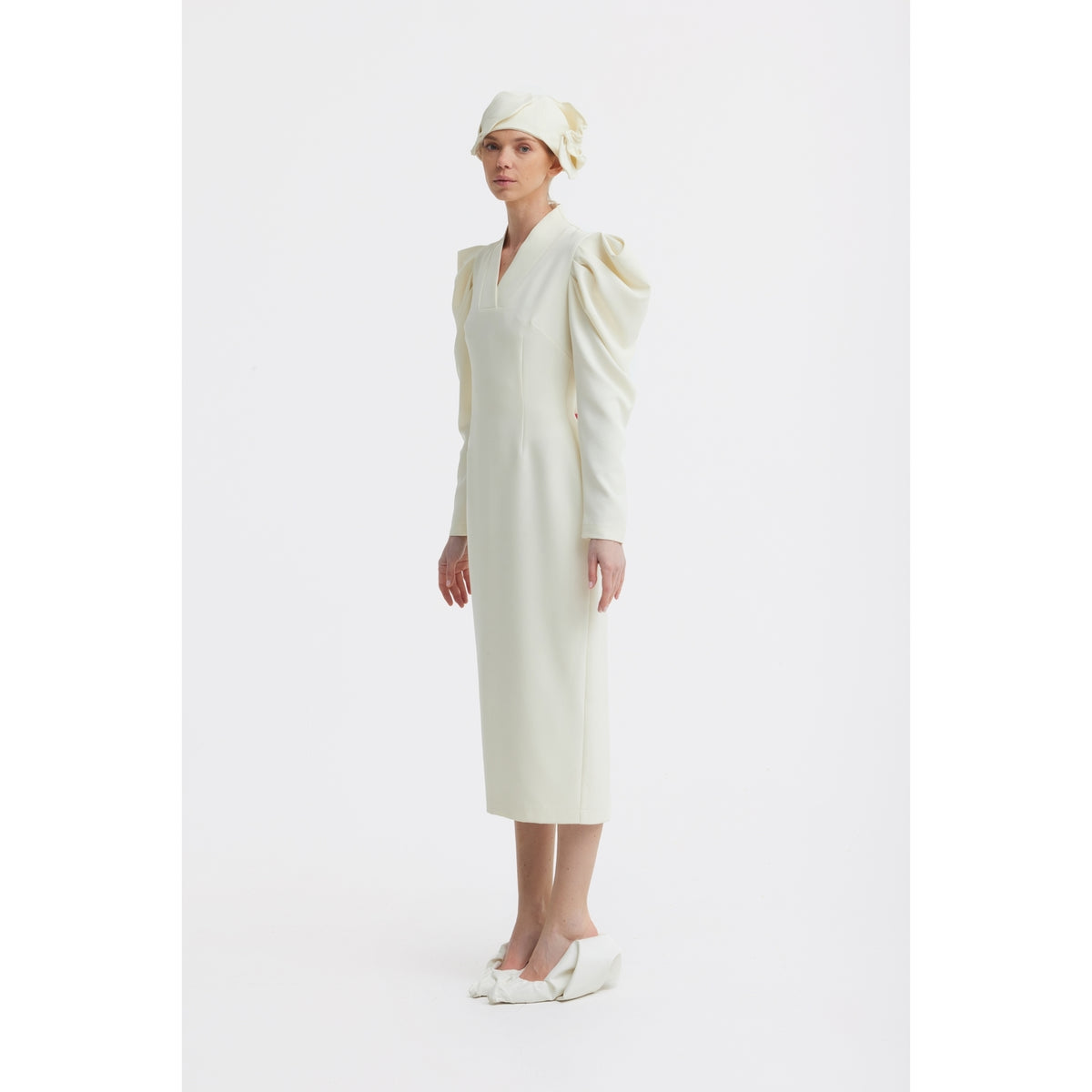 A woman stands against a white background wearing the JULIA ALLERT Crepe Midi Dress With Statement Sleeves in ivory, paired with a matching headscarf and pointed shoes adorned with large bows. She faces slightly to the side with a neutral expression.