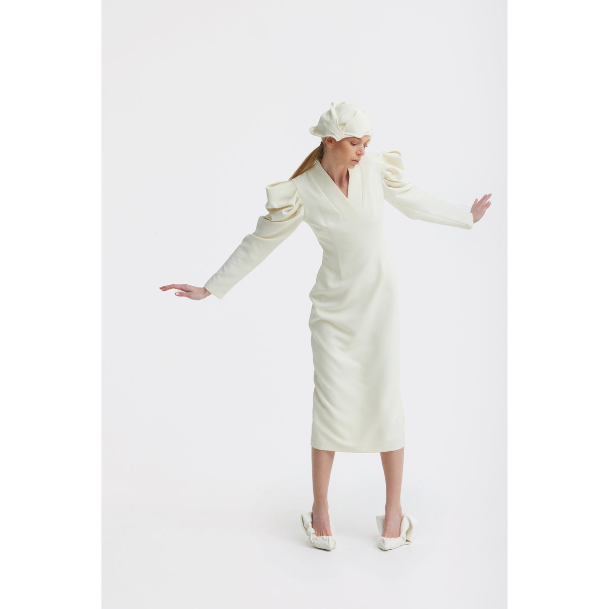 A woman models the JULIA ALLERT Crepe Midi Dress With Statement Sleeves in ivory, paired with a matching hat, posing gracefully against a plain white background with arms extended and head tilted.