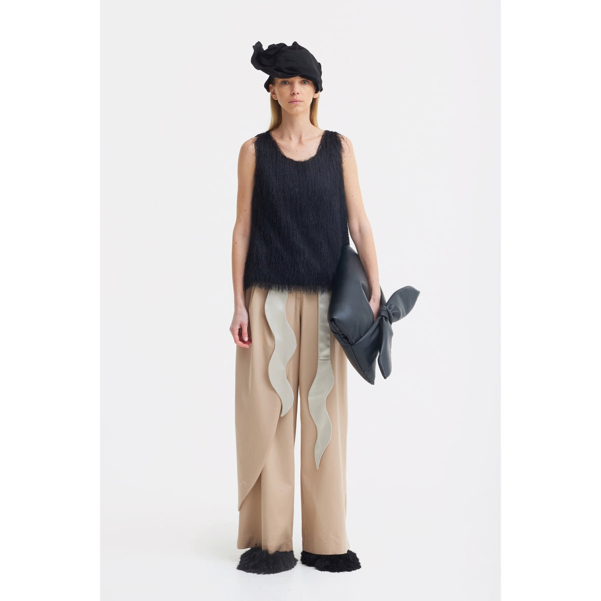 A woman stands against a white background wearing a black textured sleeveless top, JULIA ALLERT High-Waist Wrap Trousers in beige, black shoes, a black headpiece, and holding a large black bow-shaped accessory.
