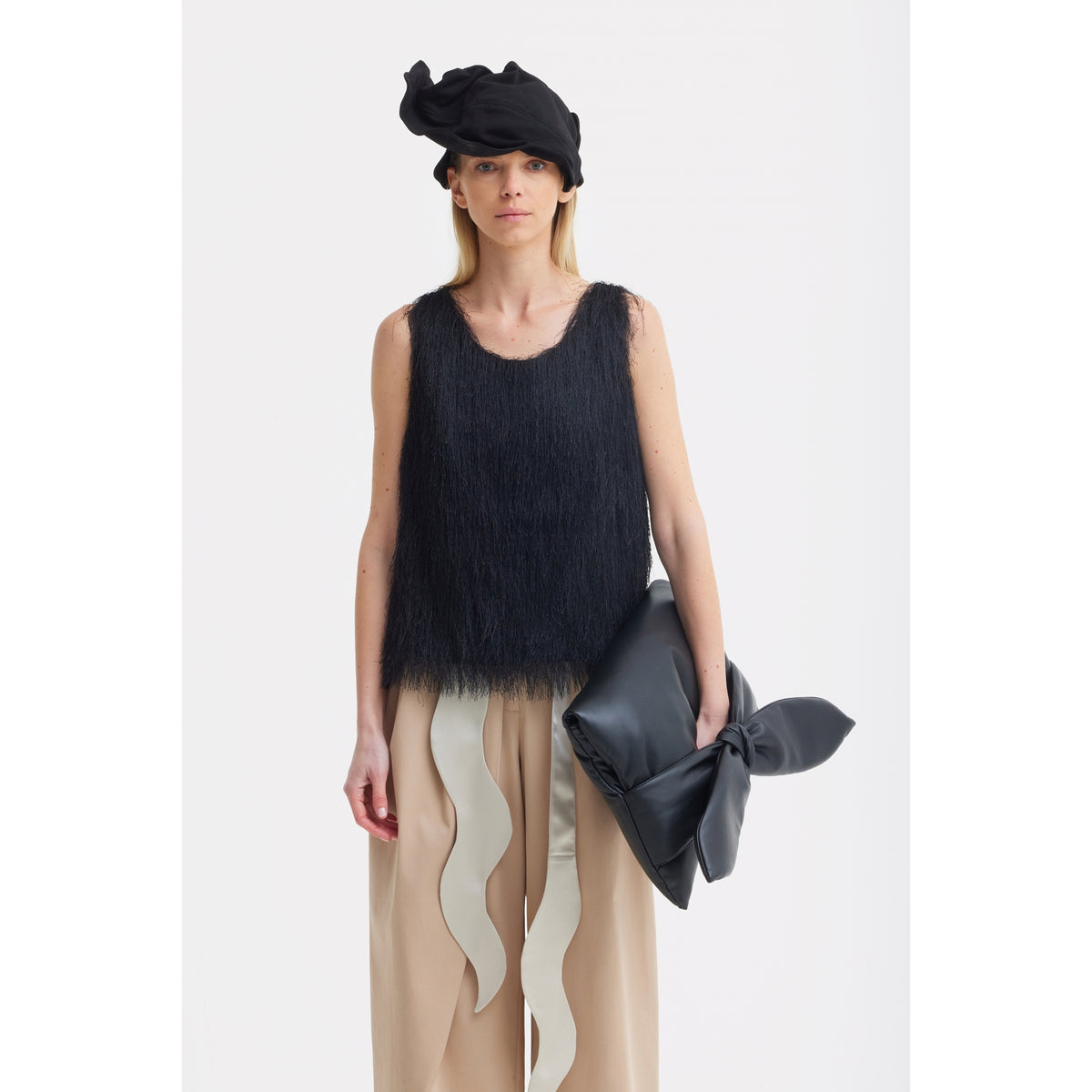 A woman wears a black textured sleeveless top, JULIA ALLERT High-Waist Wrap Trousers in beige, a black headwrap, and holds a large black clutch with a bow against a white background.