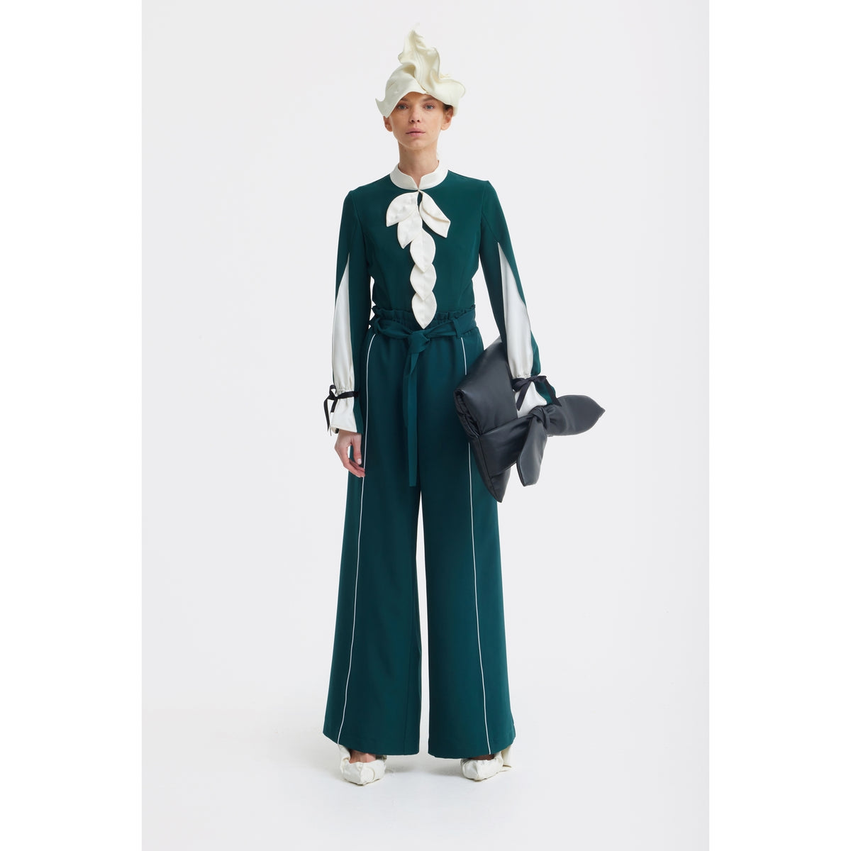 A person stands against a white background wearing the JULIA ALLERT Elegant Fitted Blouse in emerald, styled with wide-leg pants, flared sleeves, a ruffled hat, and a large black clutch for a chic look.