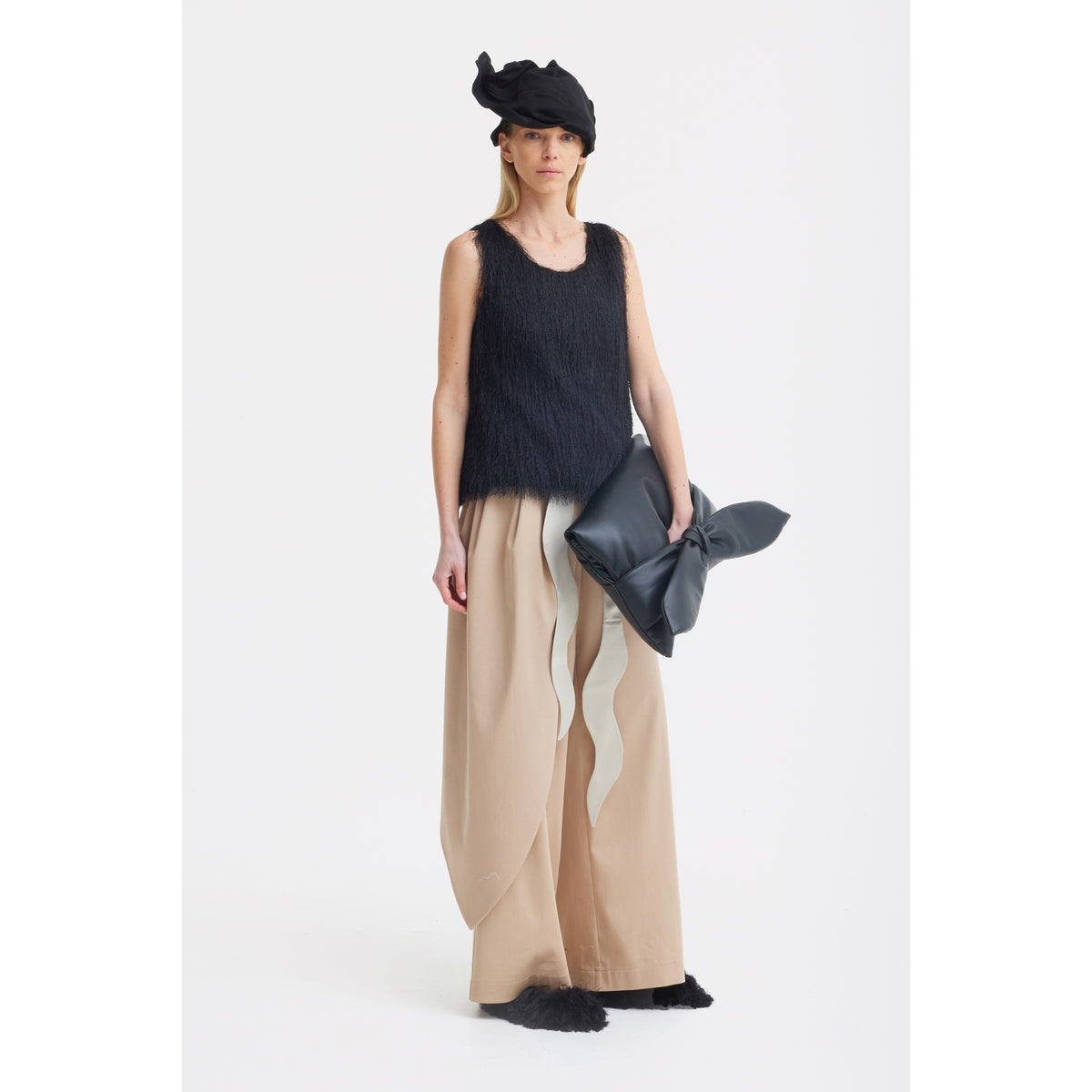 A woman stands against a white background wearing a black textured sleeveless top, JULIA ALLERT High-Waist Wrap Trousers in beige, black fuzzy shoes, a black hat, and holding a large black clutch.