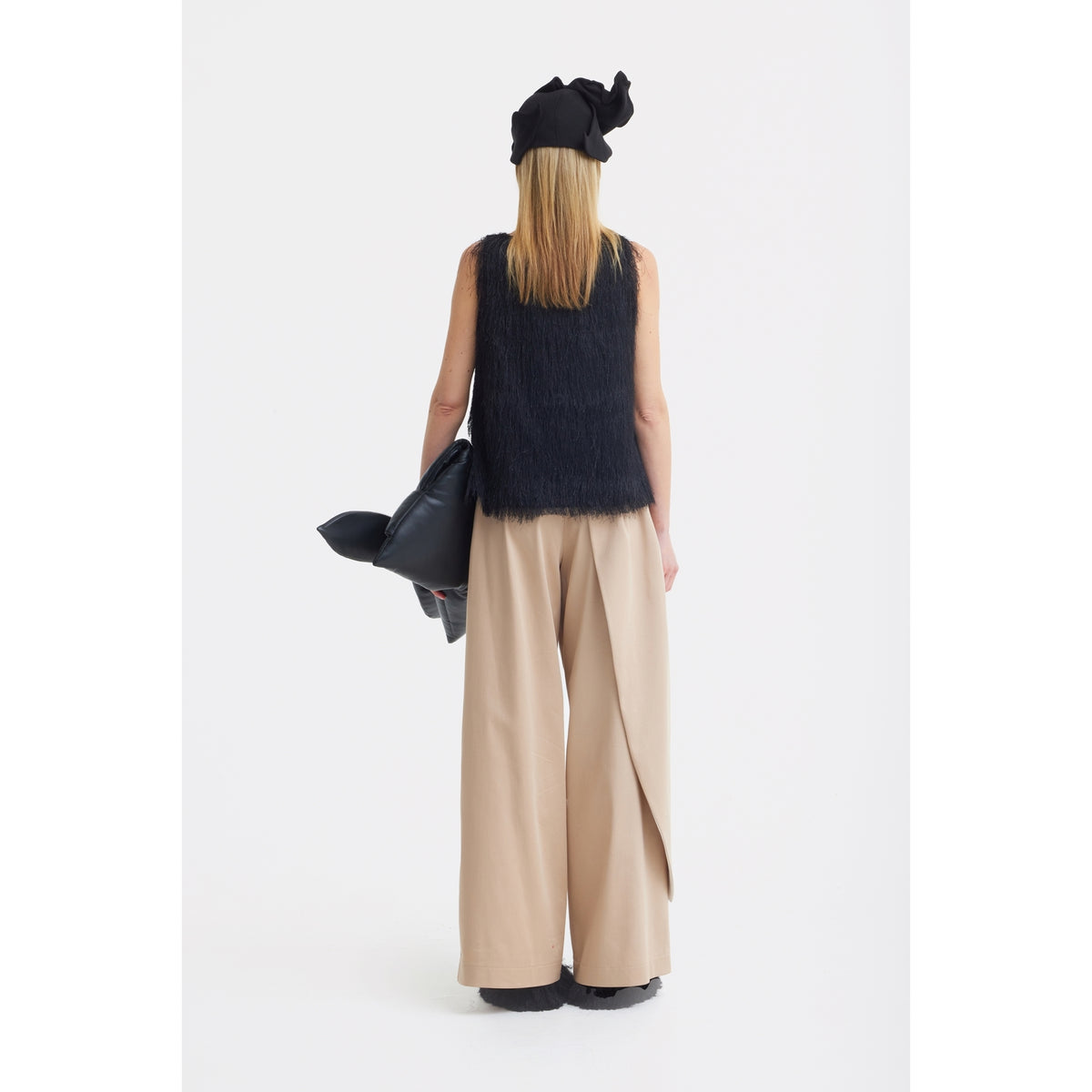 A person stands facing away against a plain white background, wearing a sleeveless black top, JULIA ALLERT High-Waist Wrap Trousers in beige, black shoes, a black hat, and holding a large black bag.