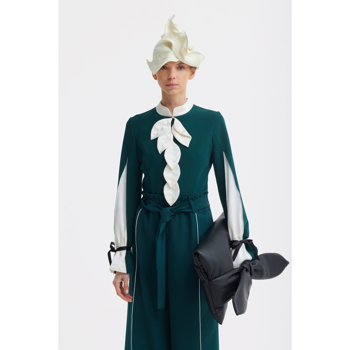 A person wears the JULIA ALLERT Elegant Fitted Blouse in emerald with white leaf details, styled with a sculptural white hat and a large black knotted clutch, against a plain white background.