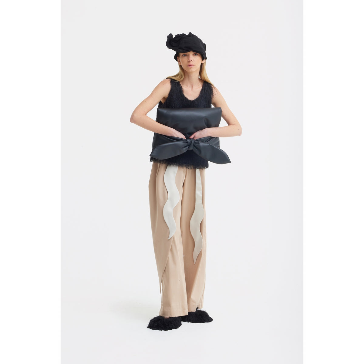 A woman in a black furry tank top, JULIA ALLERT High-Waist Wrap Trousers in beige, black furry shoes, and a black hat stands holding a large black bow against a plain white background.