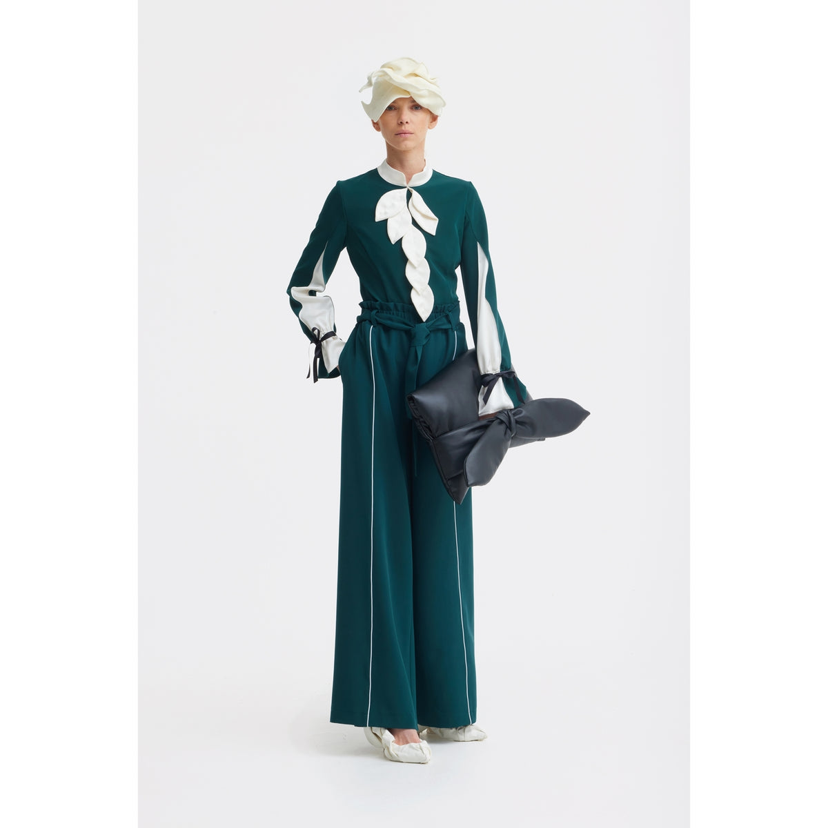 Against a white background, a person wears the JULIA ALLERT Elegant Fitted Blouse in emerald, styled with white heels, a large cream hat, and carries a large black bow-shaped bag.