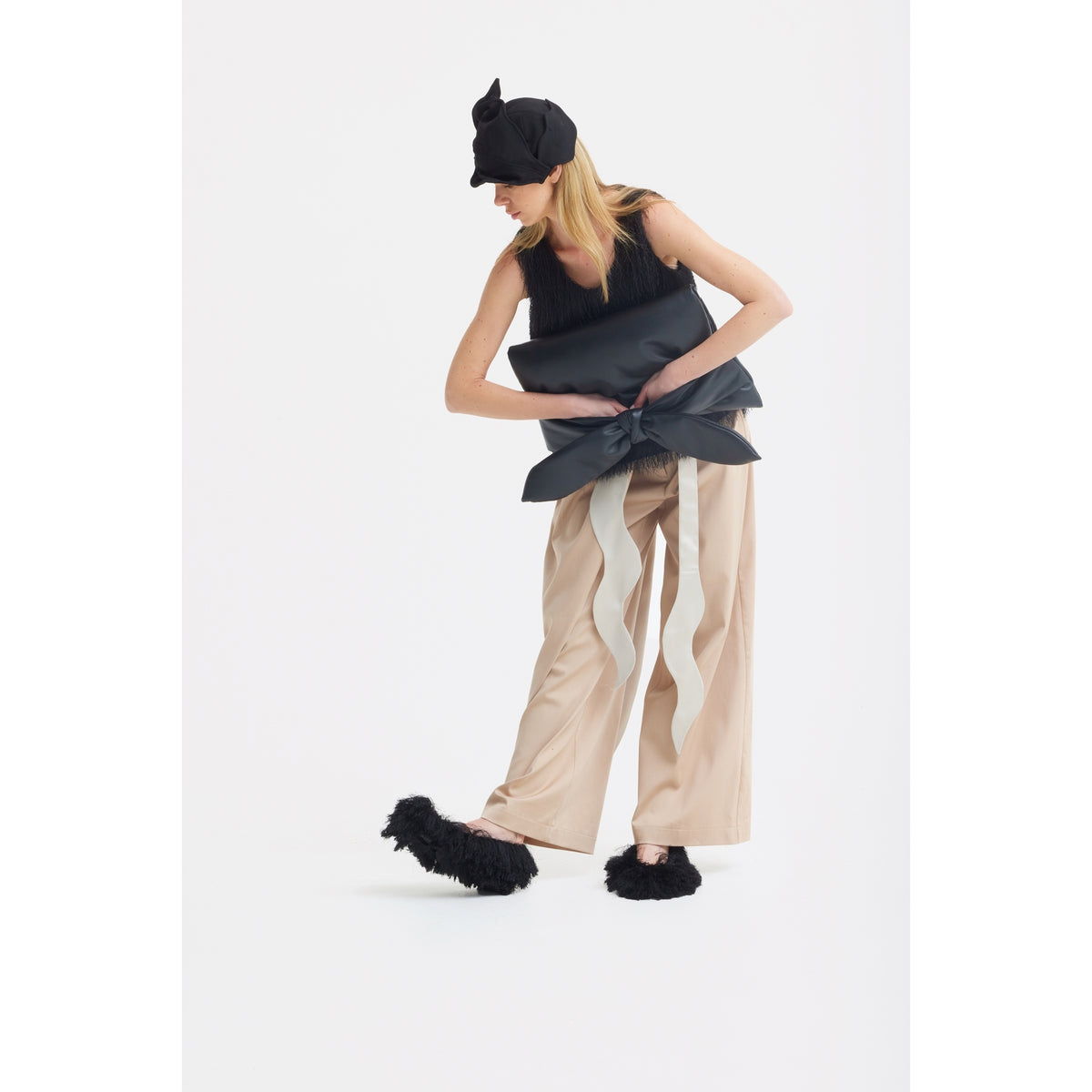 A woman in a black sleeveless top, JULIA ALLERT High-Waist Wrap Trousers in beige, black fluffy slippers, and a black hat poses holding a large black pillow-like accessory against a plain white background.