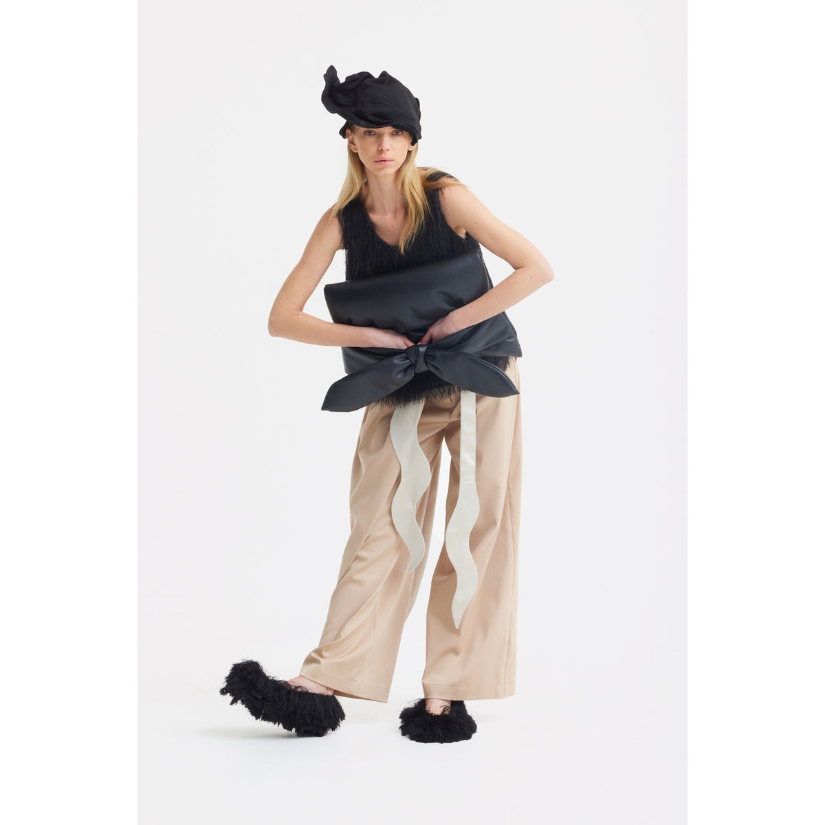 A woman wearing JULIA ALLERT High-Waist Wrap Trousers in beige, a sleeveless black top, a large fabric waist bow, black fuzzy slippers, and a sculptural black headpiece poses against a plain white background.