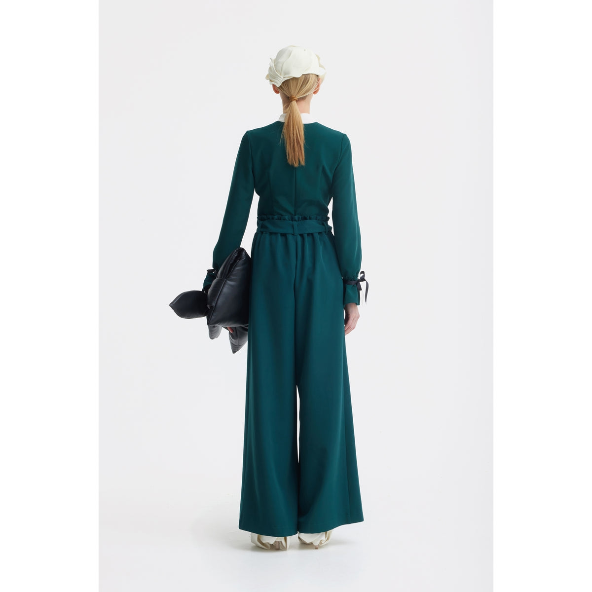 A person with long blonde hair in a ponytail wears the JULIA ALLERT Elegant Fitted Blouse in emerald, paired with a white hat and cream shoes, holding a large black bag against a plain white background.