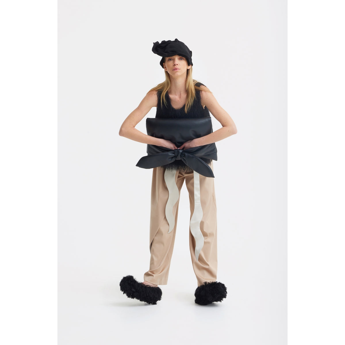 A person wearing a black sleeveless top, large black bow, JULIA ALLERT High-Waist Wrap Trousers in beige, black fuzzy slippers, and a ruffled black hat stands against a plain white background.