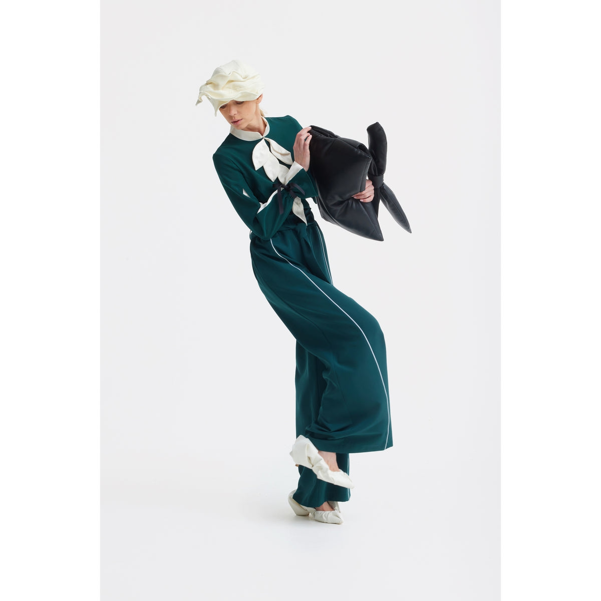 A person wearing the Elegant Fitted Blouse - Emerald by JULIA ALLERT poses against a white background, accessorized with a matching hat and holding a large black bow-shaped object.