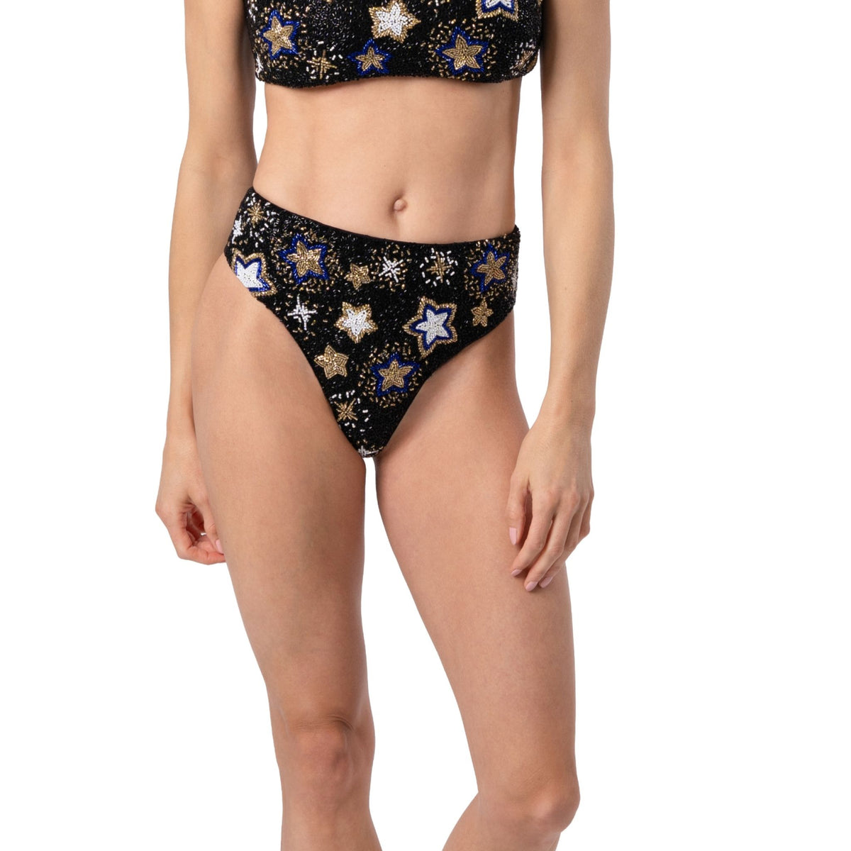 A woman models the SELF-ADORE Celeste Hand-Beaded Embellished Bikini Bottom in black, featuring gold, white, and blue star designs; her midsection and upper thighs highlight the stunning swimwear.
