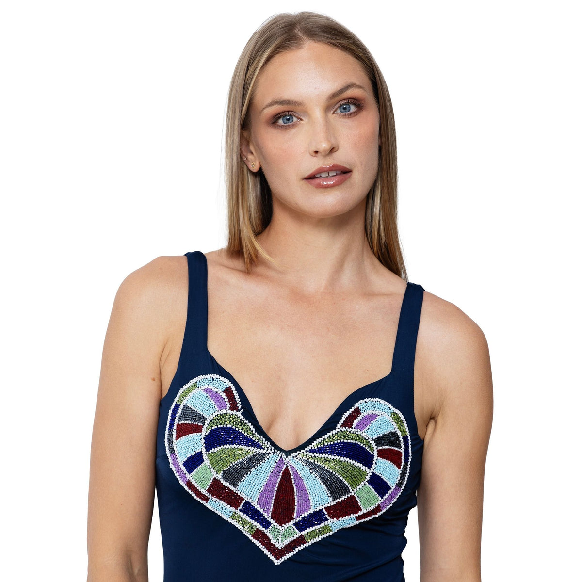 A woman with straight blonde hair wears the SELF-ADORE Amor Hand-Beaded Embellished One-Piece Swimsuit in navy, featuring a colorful heart design on the chest. She poses front-facing with a neutral expression against a white background.
