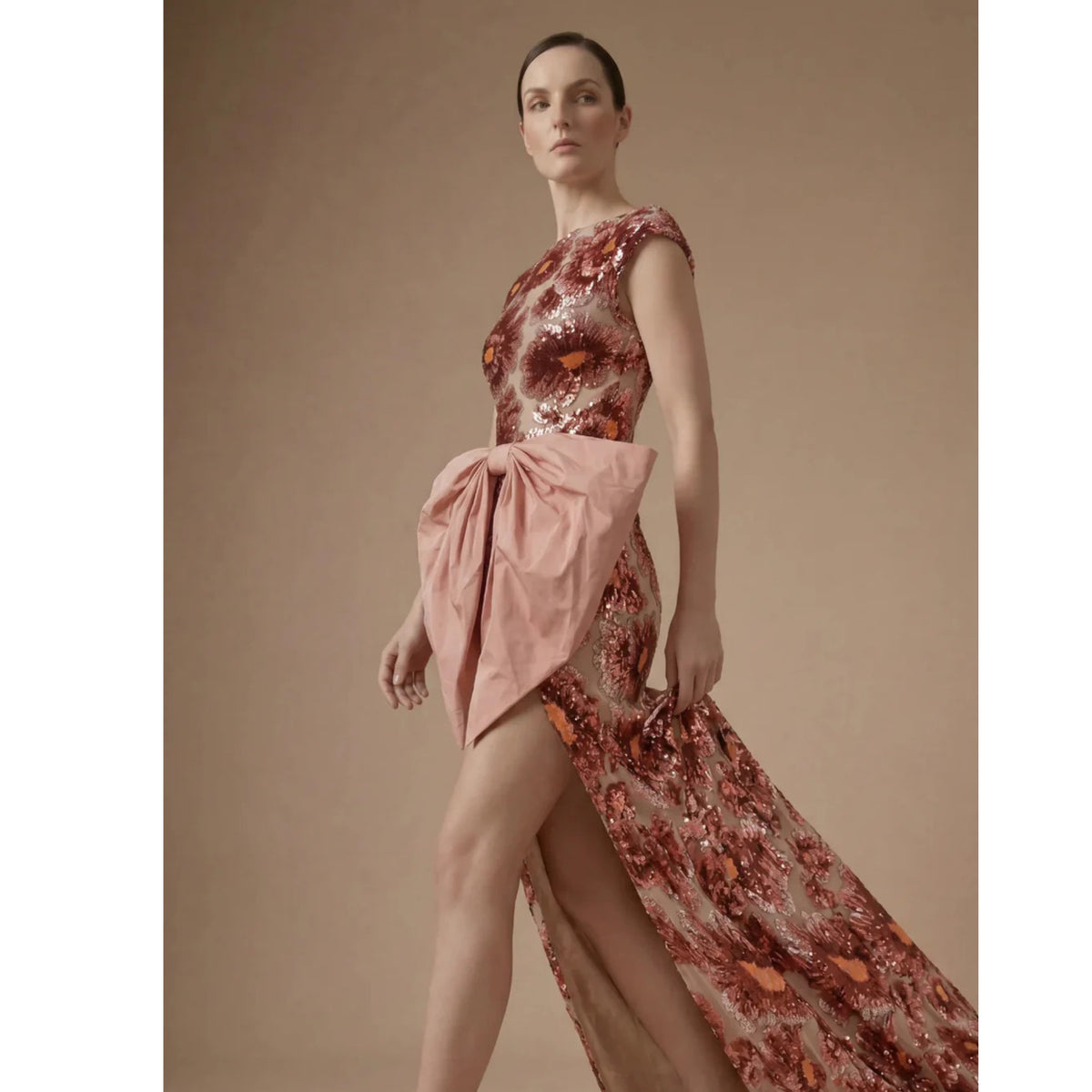 A woman models the ASHLEY STAMBOULI Coralia Dress | PR Sample, a high-slit evening gown with floral pattern and a large pink waist bow, standing confidently against a beige background.