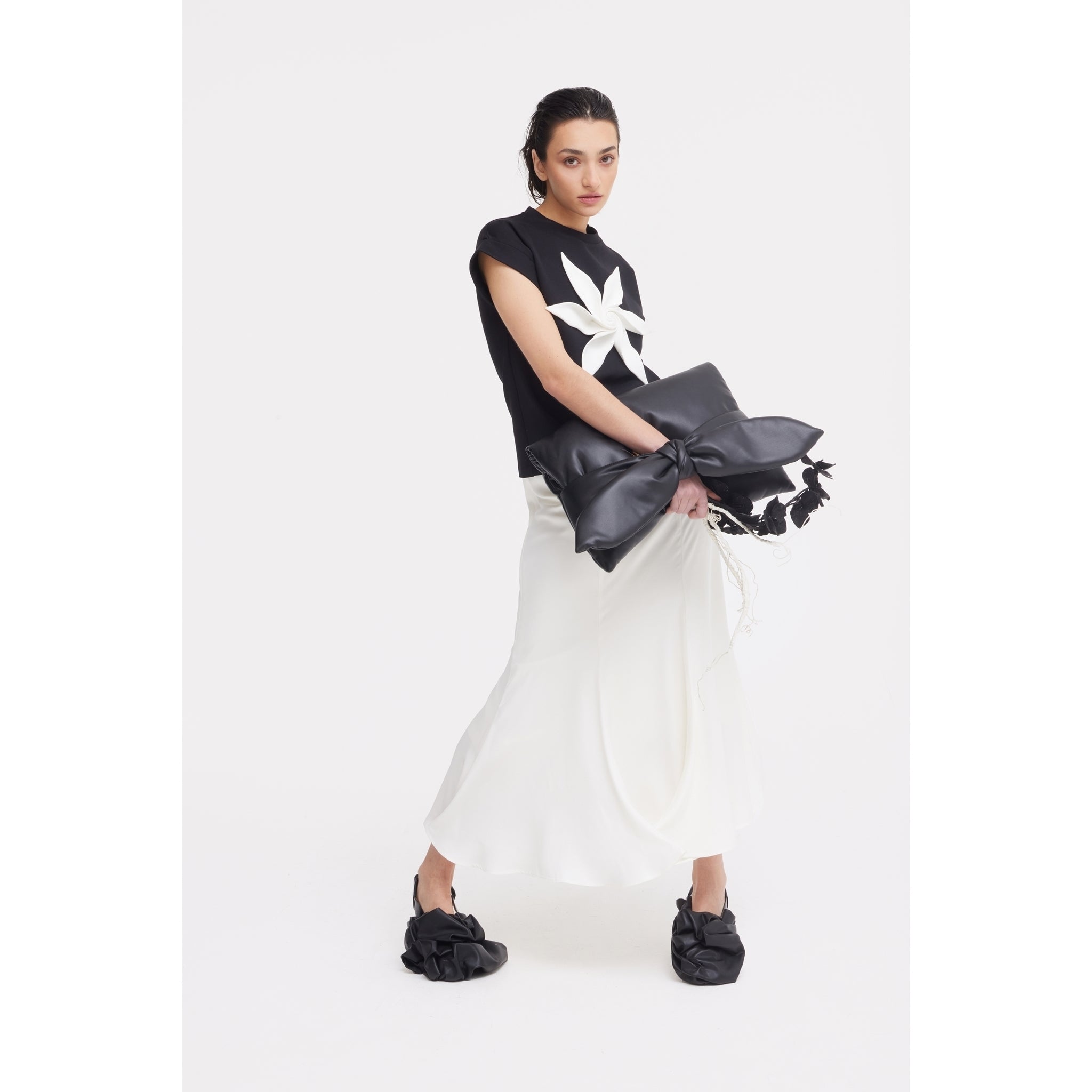 A woman poses before a white backdrop in JULIA ALLERT eveningwear: a black top with a large white flower, High Waist Double-Sided Satin Maxi Skirt - Ivory, black slip-on shoes, and a bold black clutch with an oversized bow.