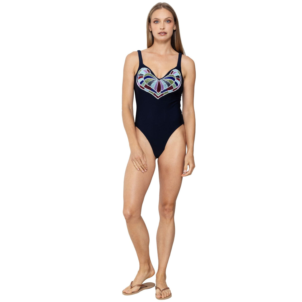 A woman models the SELF-ADORE Amor Hand-Beaded Embellished One-Piece Swimsuit in navy, featuring a vibrant chest design. She is barefoot with beige sandals and poses against a plain white background.