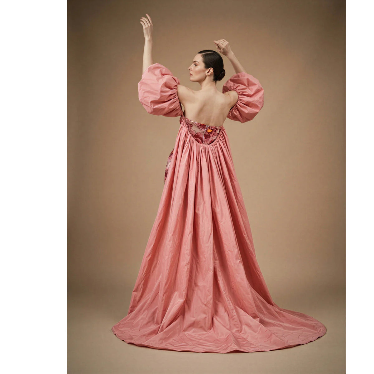 A woman stands with arms raised in the ASHLEY STAMBOULI Kaspia Gown Dress (PR Sample), a dramatic pink embellished dress with voluminous puff sleeves, low back, and a long skirt pooling on the floor against a neutral background.