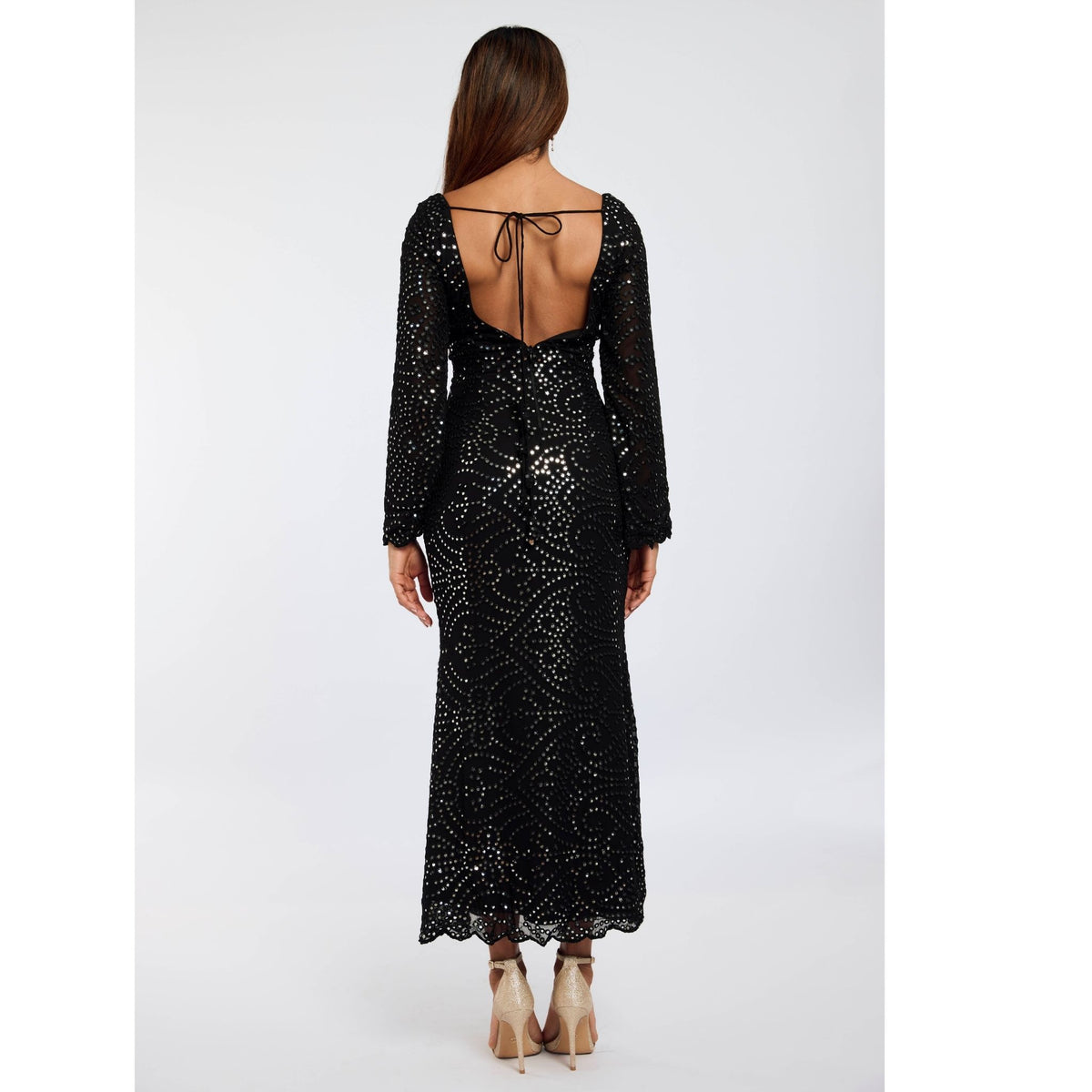 A woman with long brown hair wears the INTERNATIONAL APPEAL Black Patra Mermaid Maxi Dress | PR Sample, a luxury black maxi with sequins, open back, and tie detail, paired with beige heels against a plain white background.