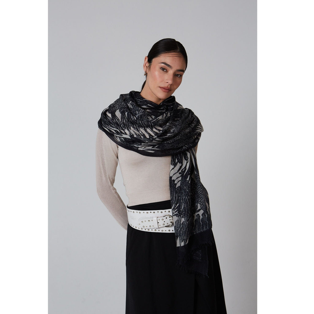 Woman with dark hair tied back wears a light beige top, black skirt, wide white belt with silver studs, and the OO-TO Black Ella Monochrome Stole—an ethical luxury cashmere scarf with a white abstract pattern—against a plain light background.