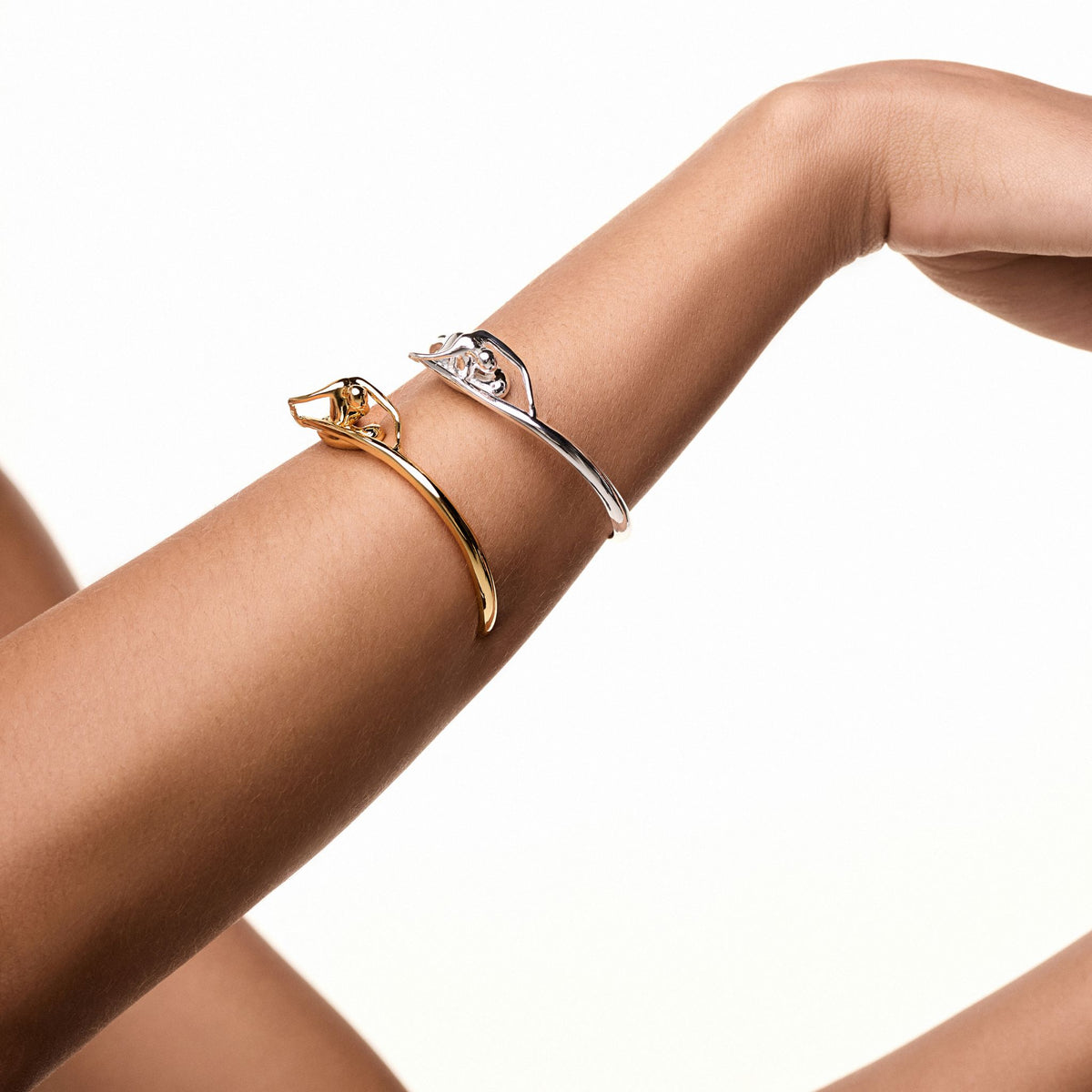 Two forearms with hands raised, each wearing an ANNA PRATA women's elegant bracelet from the Corpo E Alma collection—one Gold Instinto Bracelet and one in silver with abstract knot design—set against a plain white background.
