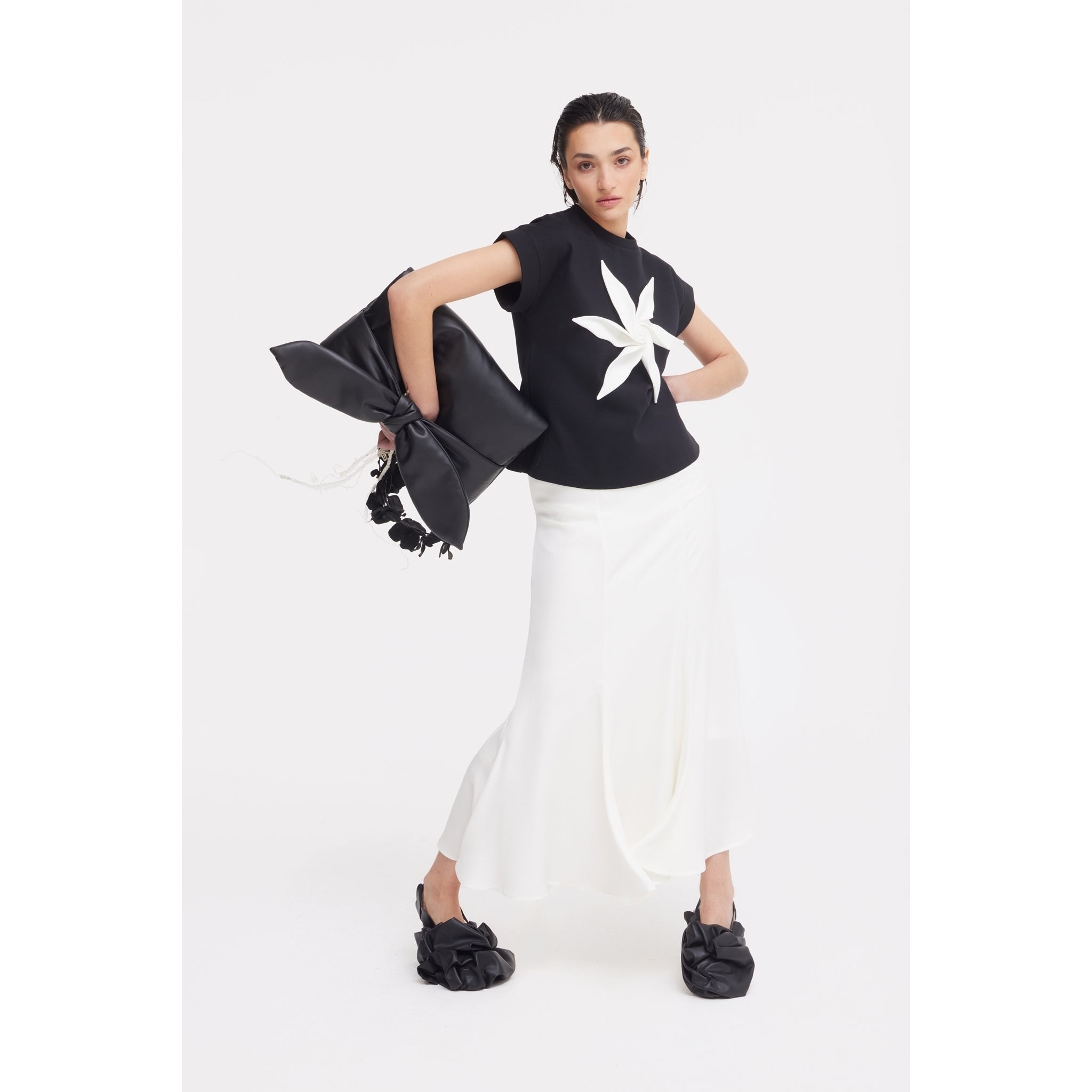 A woman poses in JULIA ALLERT eveningwear: a black top with a white star-shaped flower, the High Waist Double-Sided Satin Maxi Skirt – Ivory, and black shoes with oversized bows, holding a large black bow accessory against a plain white background.