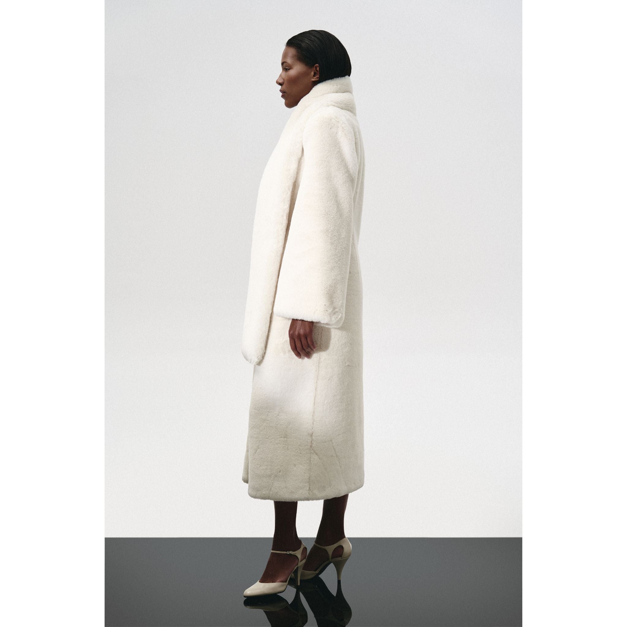 A person in profile wears the KRIS MARÁN Fur Maxi Coat With Shawl Collar—long, white faux fur with wide sleeves—paired with cream heels against a plain light background.