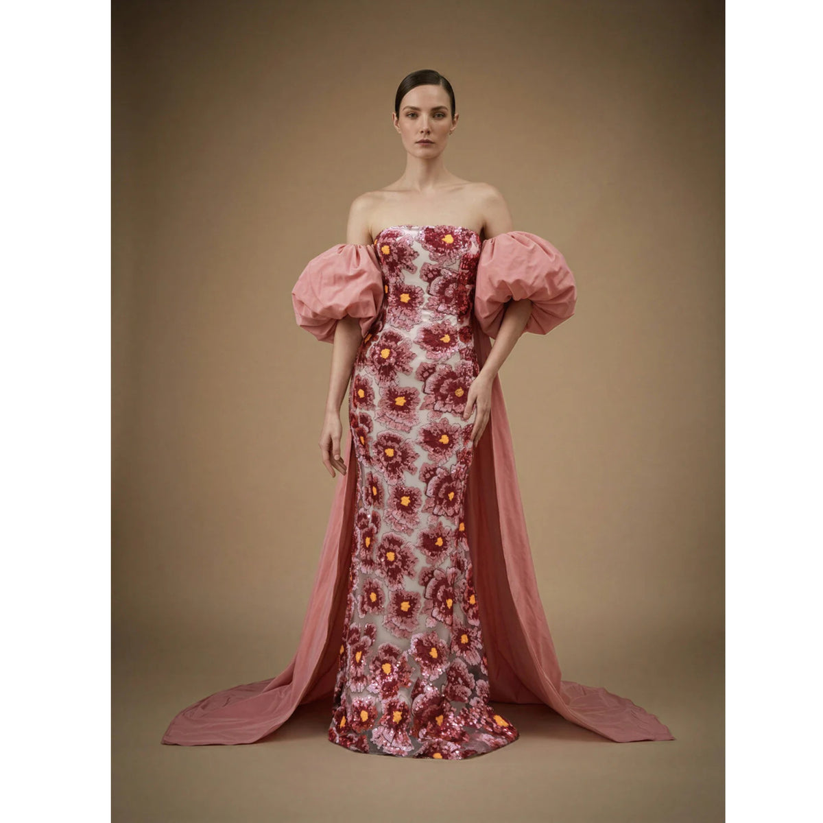 A woman poses against a plain background wearing the ASHLEY STAMBOULI Kaspia Gown Dress | PR Sample, featuring large pink puff sleeves, a floral bodice, and an elegant pink train flowing behind her.