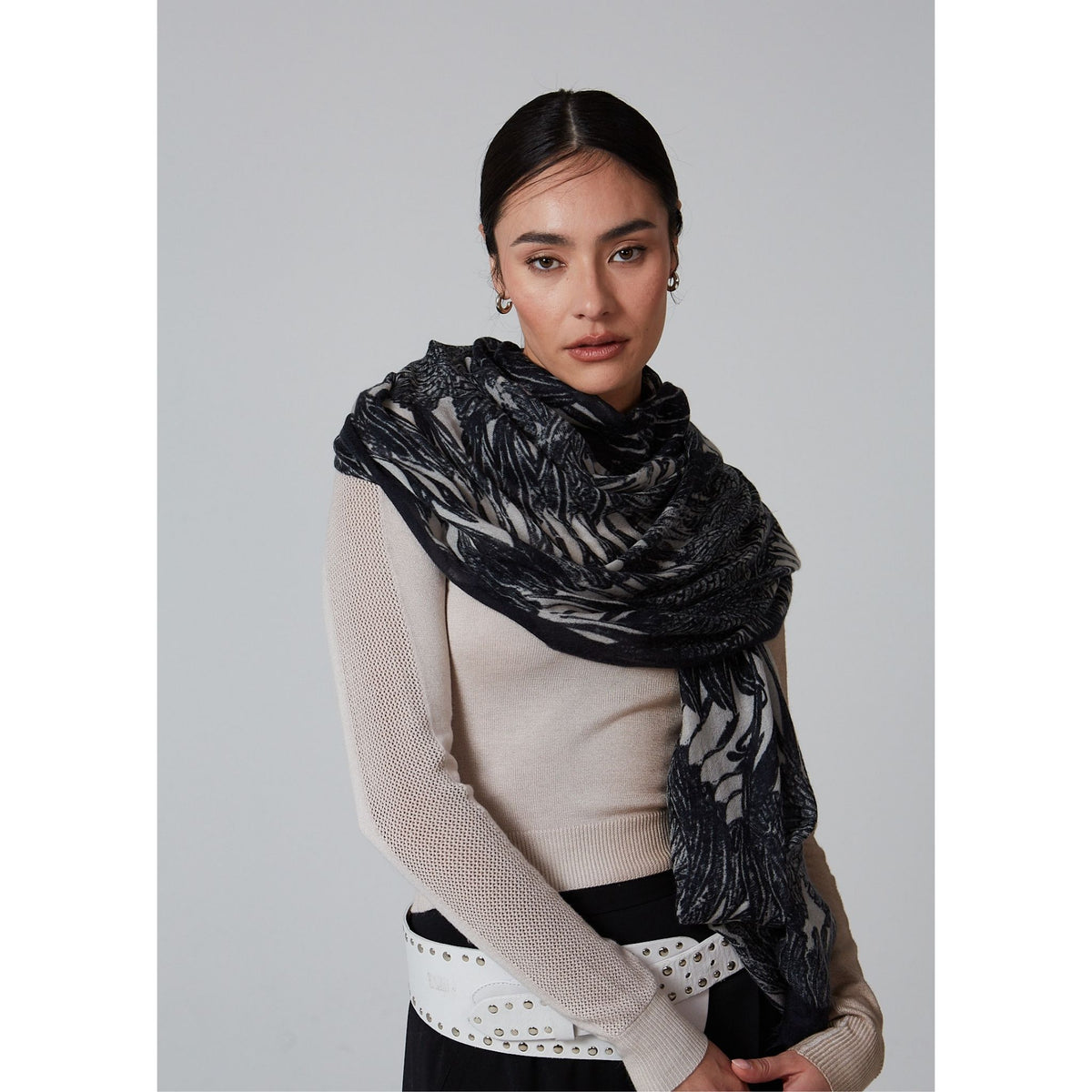 A woman with dark hair tied back wears a light beige ribbed sweater, the OO-TO Black Ella Monochrome Stole draped over her shoulders, a white belt with metal eyelets, and black pants, standing against a plain gray background.