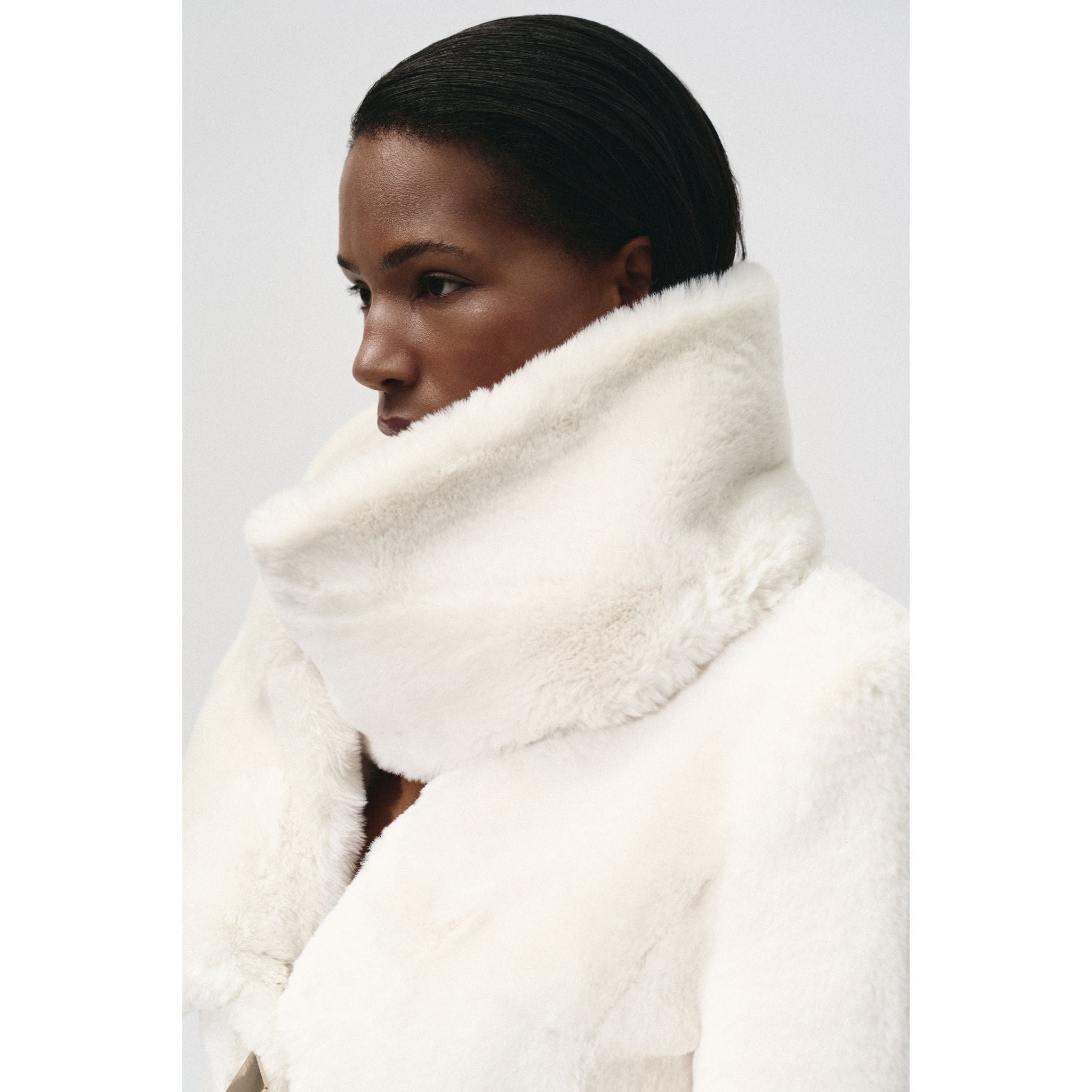A person with dark hair pulled back wears the KRIS MARÁN Fur Maxi Coat With Shawl Collar—a thick, white, fluffy faux fur coat with a high, oversized collar covering their lower face, set against a plain light background.
