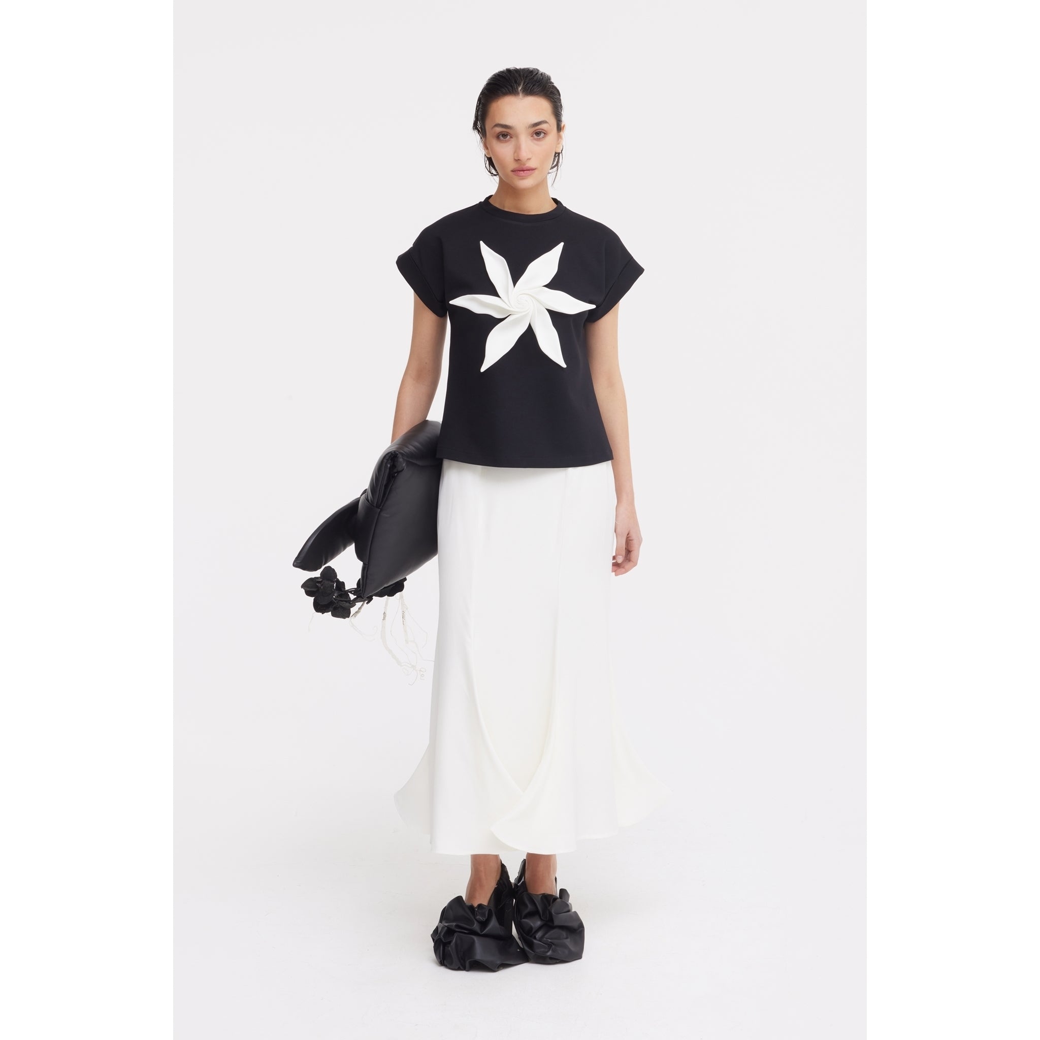 A woman poses against a white background in a JULIA ALLERT black top with a bold white flower, the High Waist Double-Sided Satin Maxi Skirt in Ivory, black ruffled shoes, and a large black floral-accented bag—an elegant evening look.