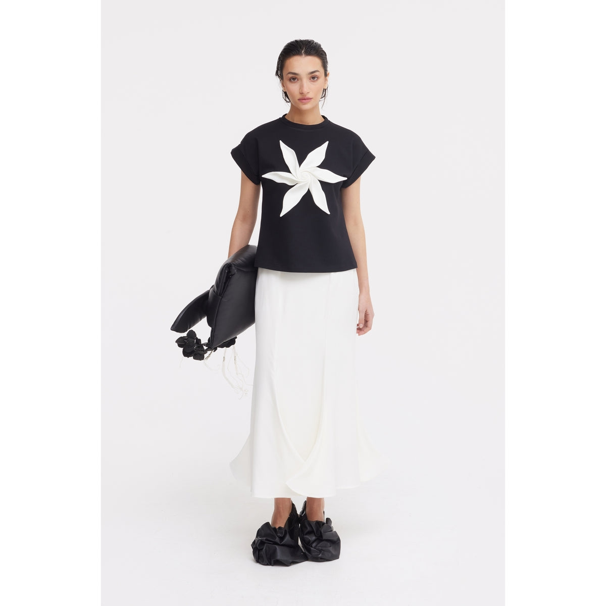 A woman poses against a white background in a JULIA ALLERT black top with a bold white flower, the High Waist Double-Sided Satin Maxi Skirt in Ivory, black ruffled shoes, and a large black floral-accented bag—an elegant evening look.