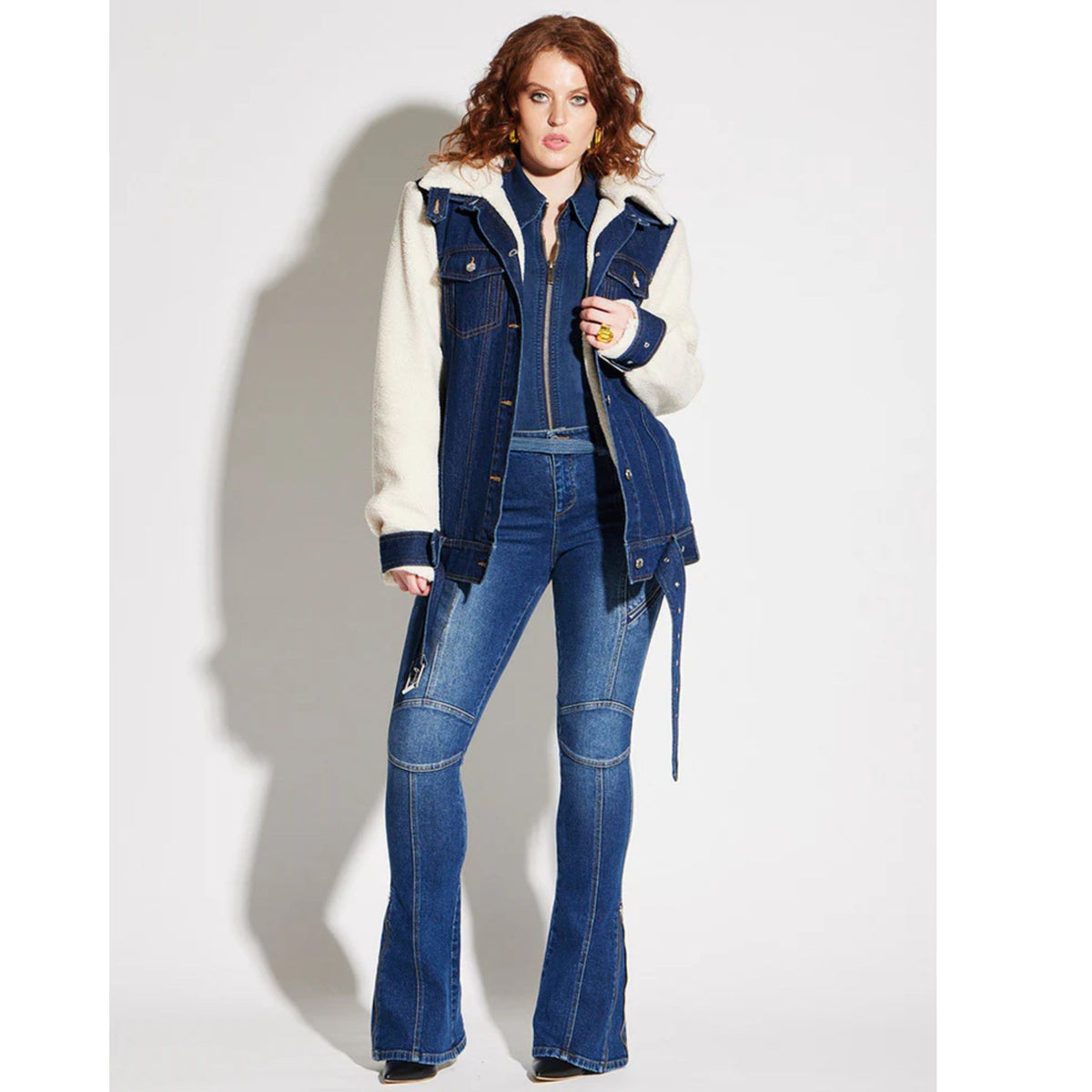 A person with curly hair poses against a white background, wearing a denim jacket with white sleeves, black top, and CIRCULAR PATH Circular Path X Love Tara Zelda Zipper Jean | PR Sample—blue flared jeans with knee patches—and black shoes.