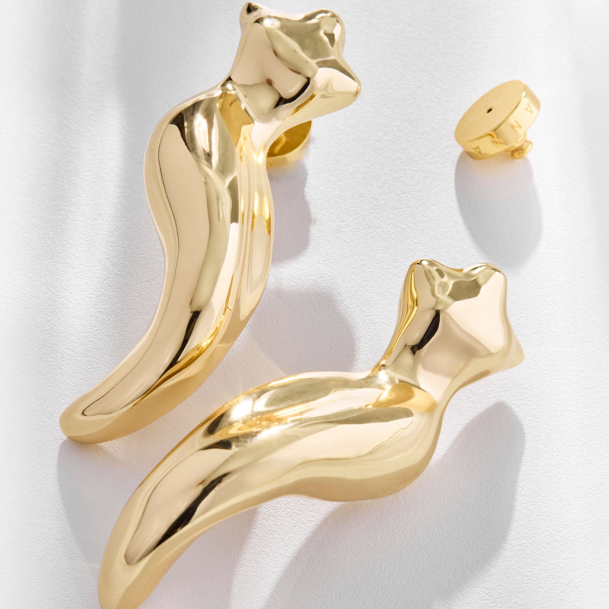 Two shiny Gold Instinto Earrings from ANNA PRATA's Corpo E Alma Collection, shaped like abstract human torsos, are displayed on a light surface with one small round earring back shown separately above the bold earrings.
