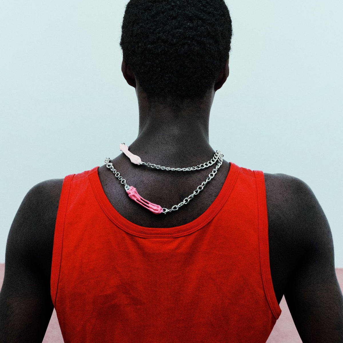 Person wearing a red tank top with two necklaces, one pink and one silver, on a light blue background.