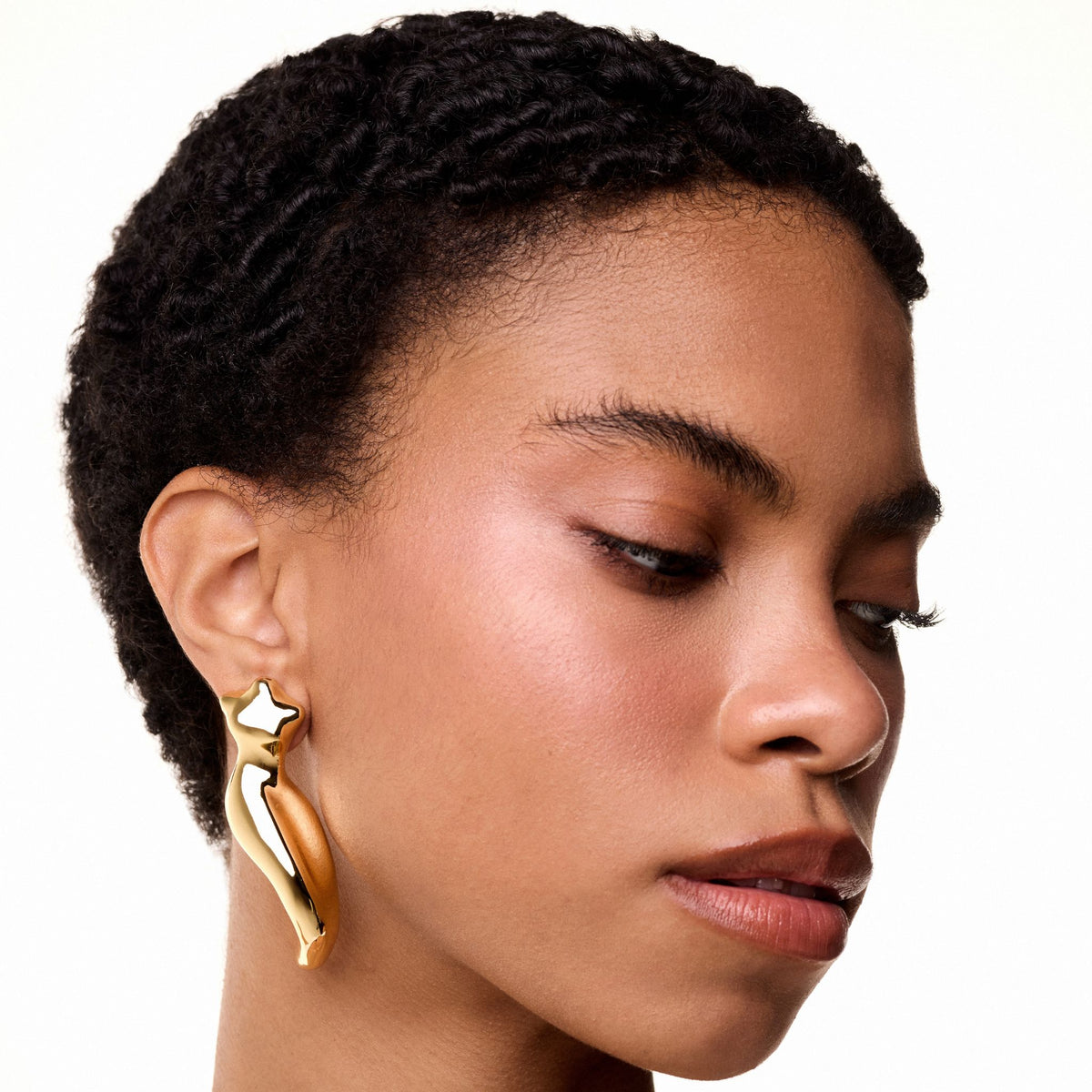 A woman with short, textured hair wears ANNA PRATA's Gold Instinto Earring from the Corpo E Alma Collection, shaped like a stylized figure with a star. Her smooth, glowing skin and calm expression are softly illuminated as she faces downward.