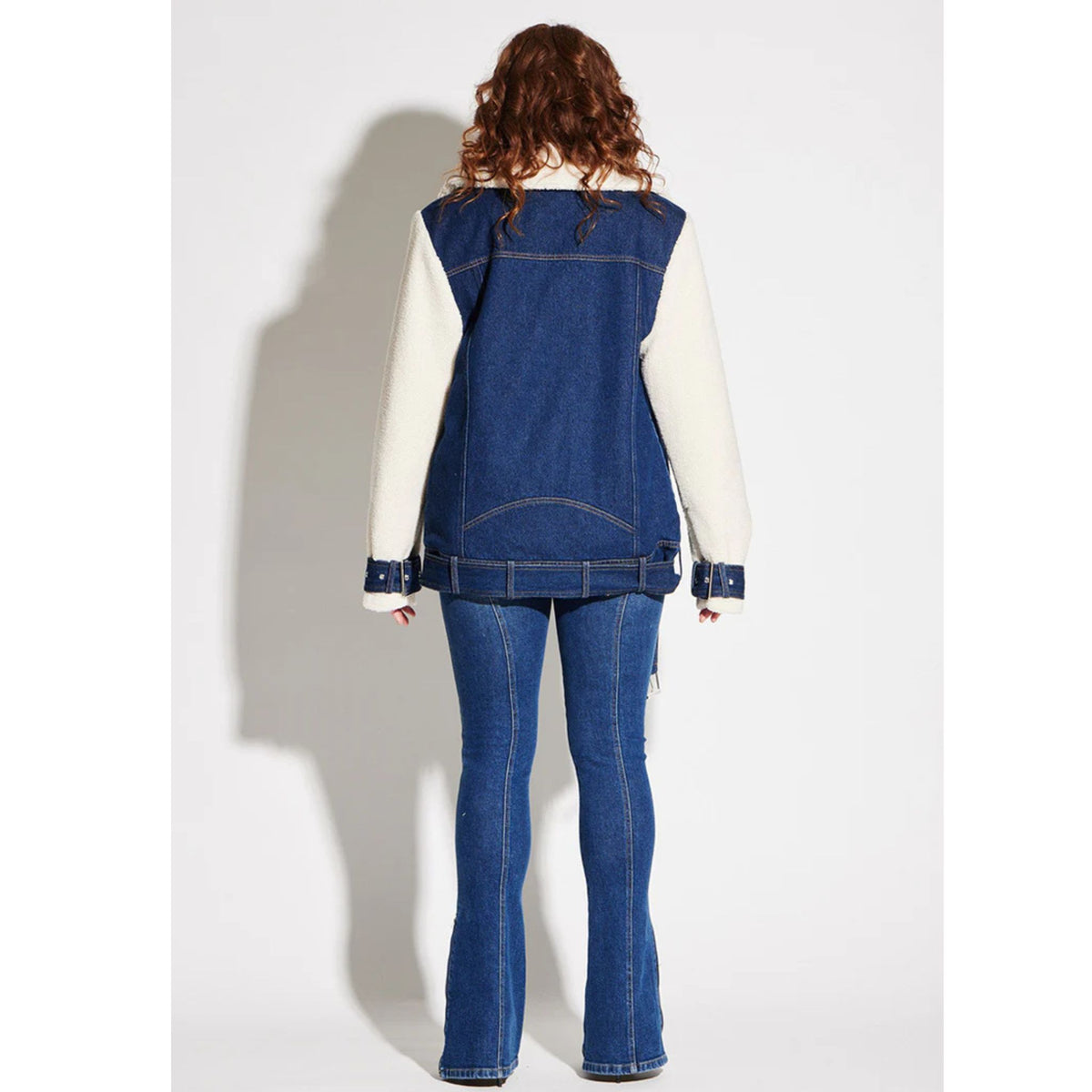 A person with curly hair is seen from behind, wearing a blue denim vest over a white long-sleeve top and the CIRCULAR PATH X Love Tara Zelda Zipper Jean | PR Sample—an eye-catching statement—against a plain white background.