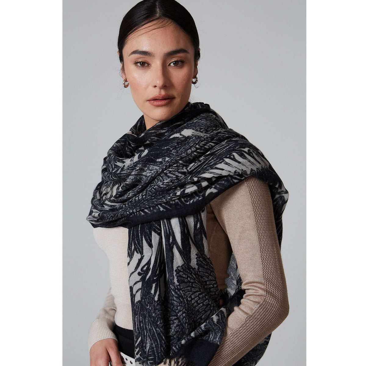 A woman with dark hair pulled back wears a beige long-sleeve top and the OO-TO Black Ella Monochrome Stole, handcrafted in Nepal, draped over her shoulders, standing against a plain light gray background.