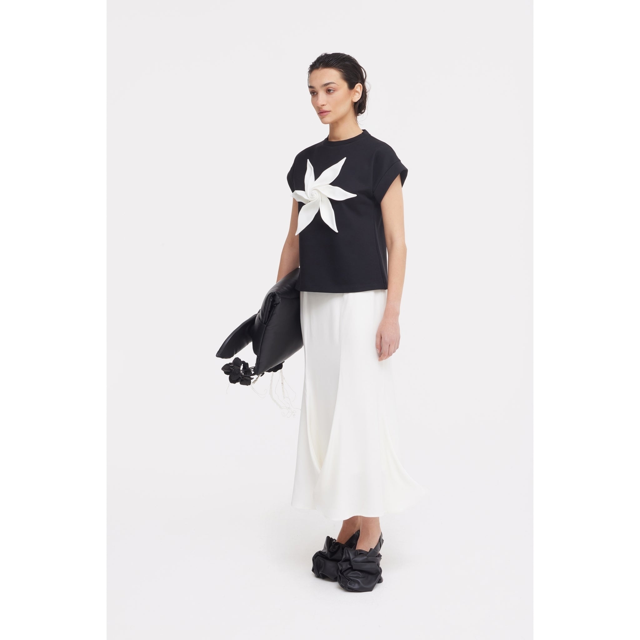 A woman stands against a white background in a black top with a star-flower design, JULIA ALLERT High Waist Double-Sided Satin Maxi Skirt in ivory, black shoes with decorative details, and a large black floral-accented bag—ideal for chic eveningwear.