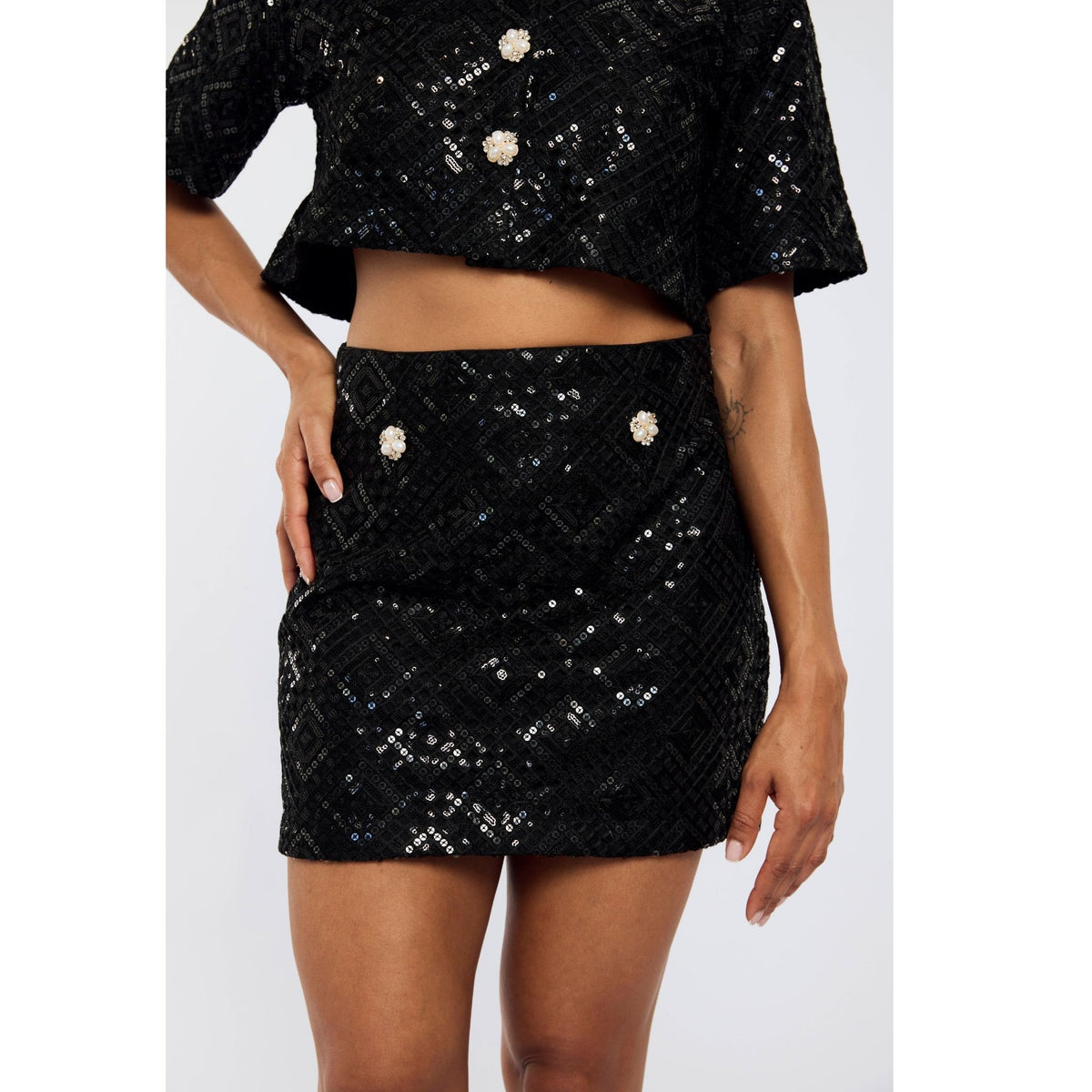 A person wears INTERNATIONAL APPEAL’s Sequin Mini Skirt | PR Sample: a black, sequined two-piece set with a crop top and high-waisted mini skirt featuring decorative buttons. Only the torso and upper legs are shown.