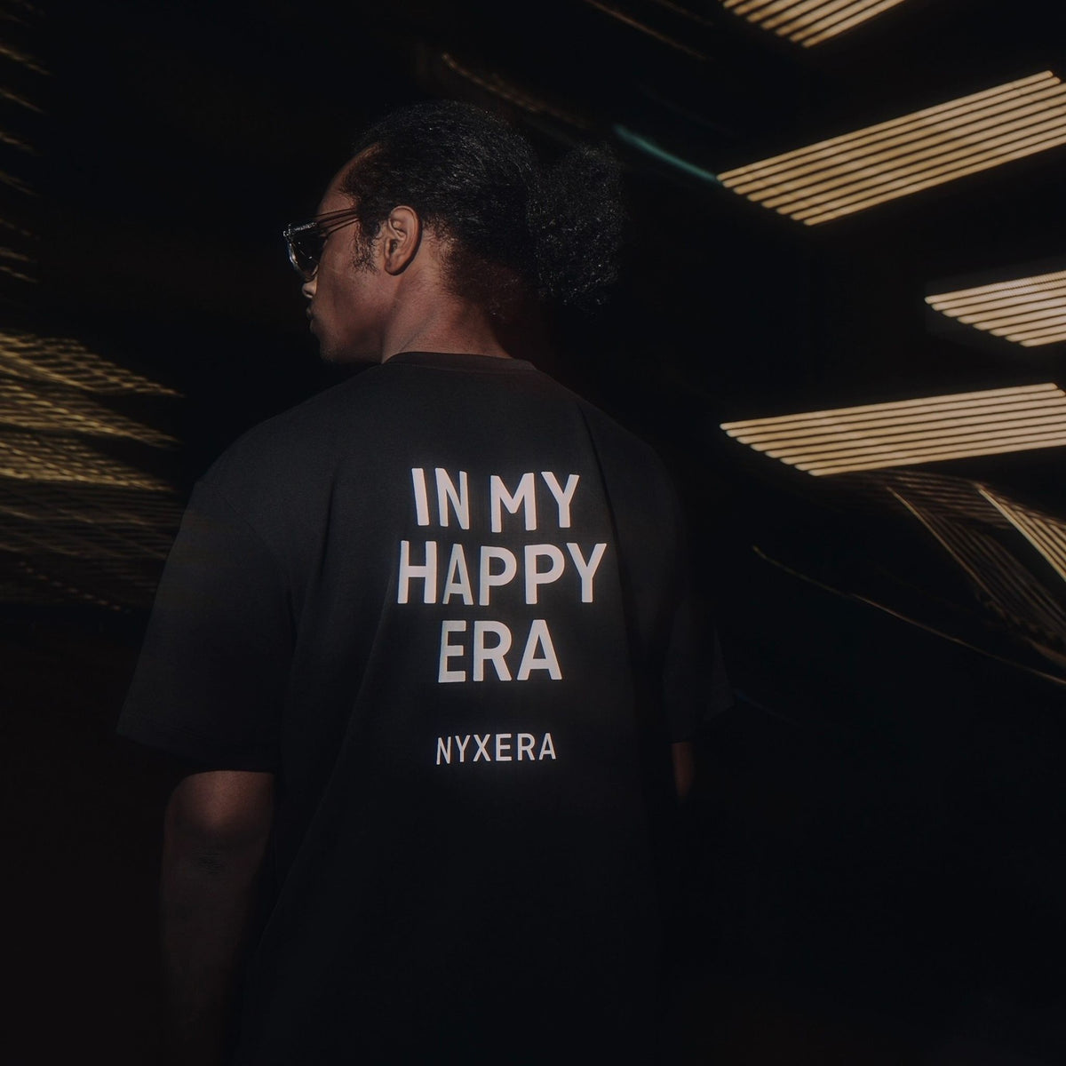 Person wearing a black t-shirt with &#39;IN MY HAPPY ERA NYXERA&#39; text on the back, standing against a dark background.