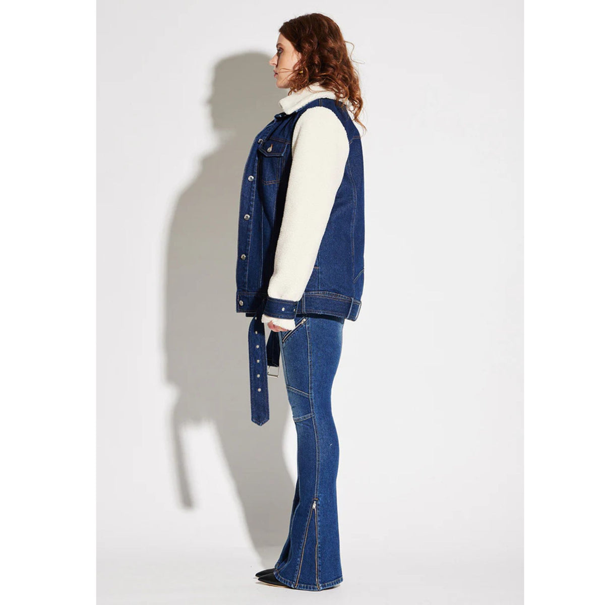 A woman with curly hair stands in profile against a white background, wearing a blue denim jacket with white sherpa sleeves and CIRCULAR PATH X Love Tara Zelda Zipper Jean (PR Sample), holding a matching denim belt for a standout look.