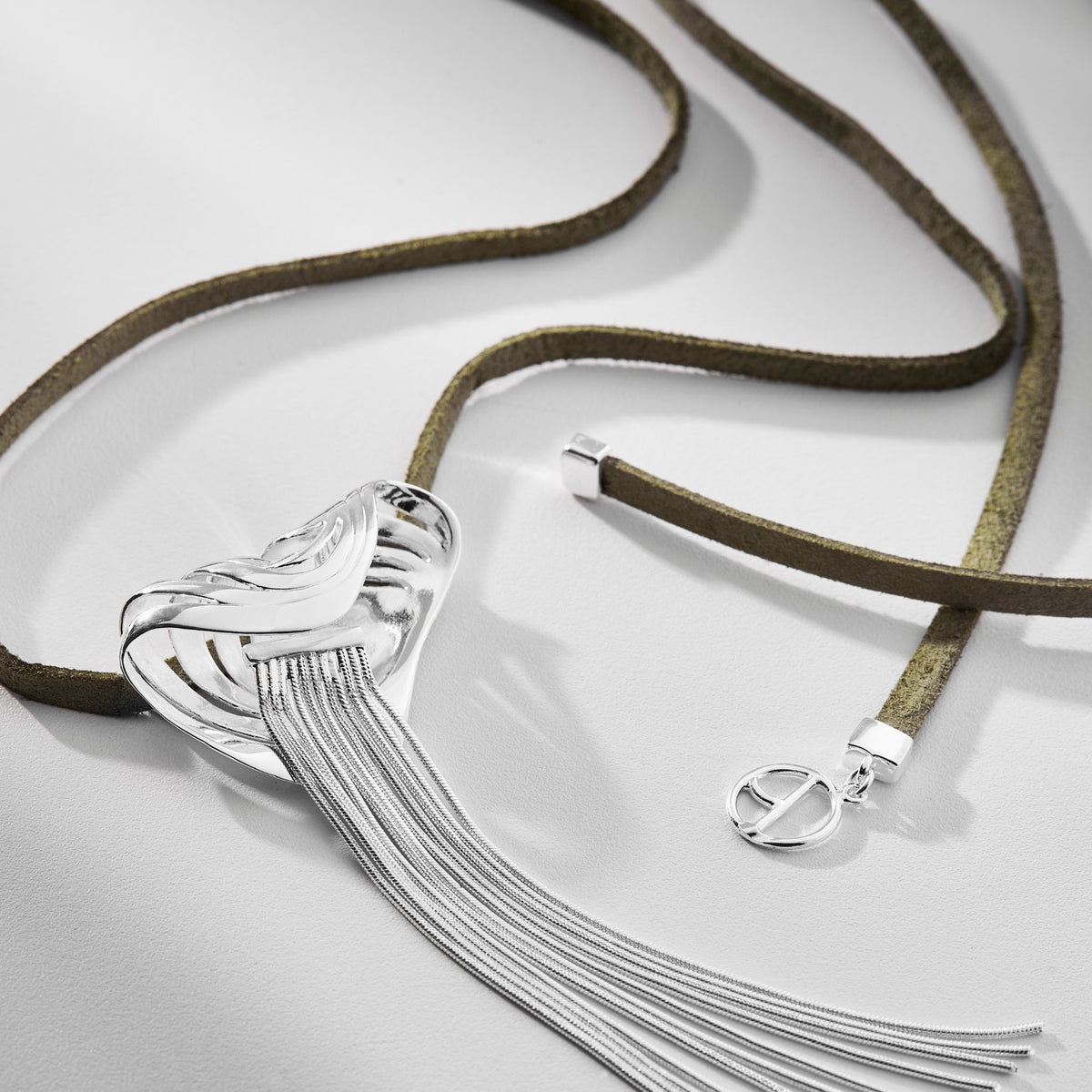 The Silver Rastro Necklace from ANNA PRATA’s Corpo E Alma Collection features a silver pendant with fringe-like strands, an olive green suede cord, and a small silver charm, all shown on a light gray surface.