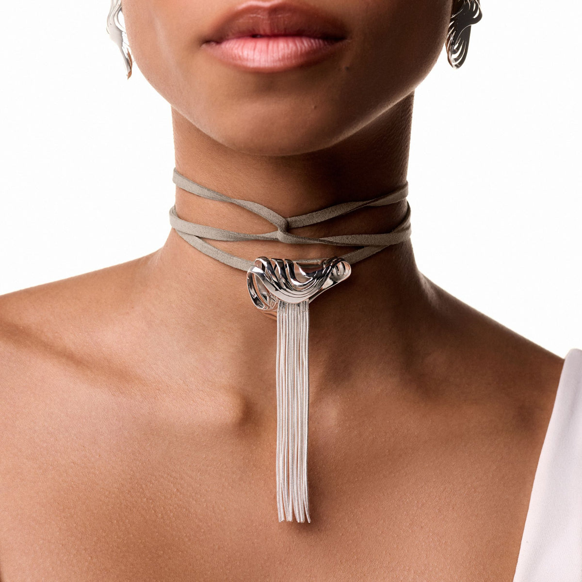 A close-up of a woman wearing the ANNA PRATA Silver Rastro Necklace from the Corpo E Alma Collection, featuring a polished metal accent and long silver fringe, paired with a white asymmetrical top.