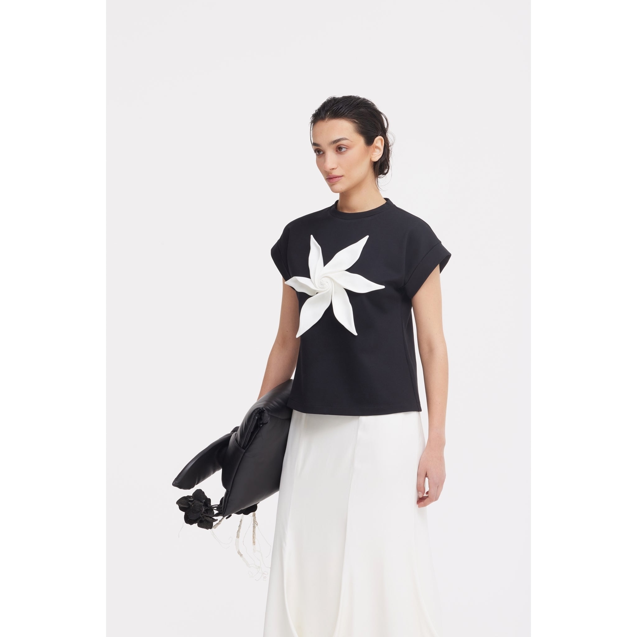 A woman in a black top with a bold white flower stands against a white background, wearing the JULIA ALLERT High Waist Double-Sided Satin Maxi Skirt in Ivory and holding a chic black bag—ideal for elegant eveningwear.