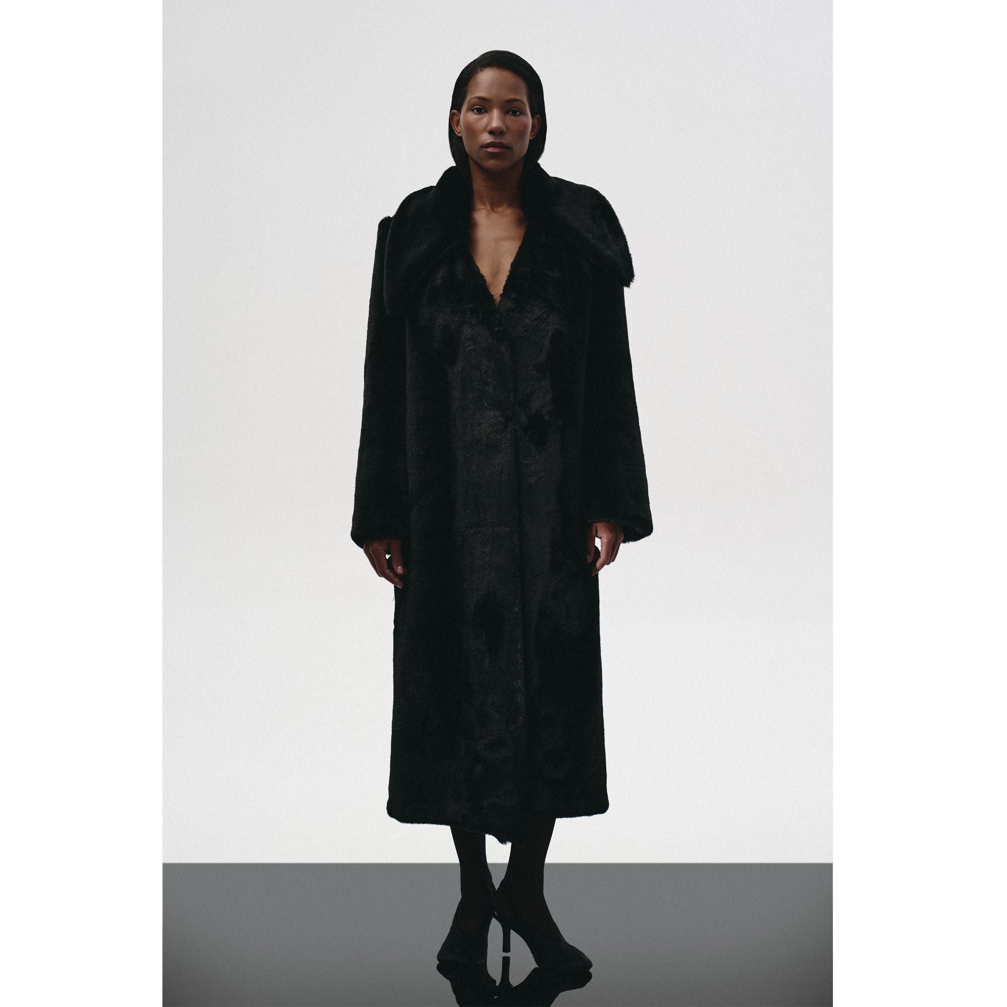 A woman with short hair faces forward against a plain white background, wearing the KRIS MARÁN Fur Maxi Coat With Oversized Collar, her arms relaxed at her sides.